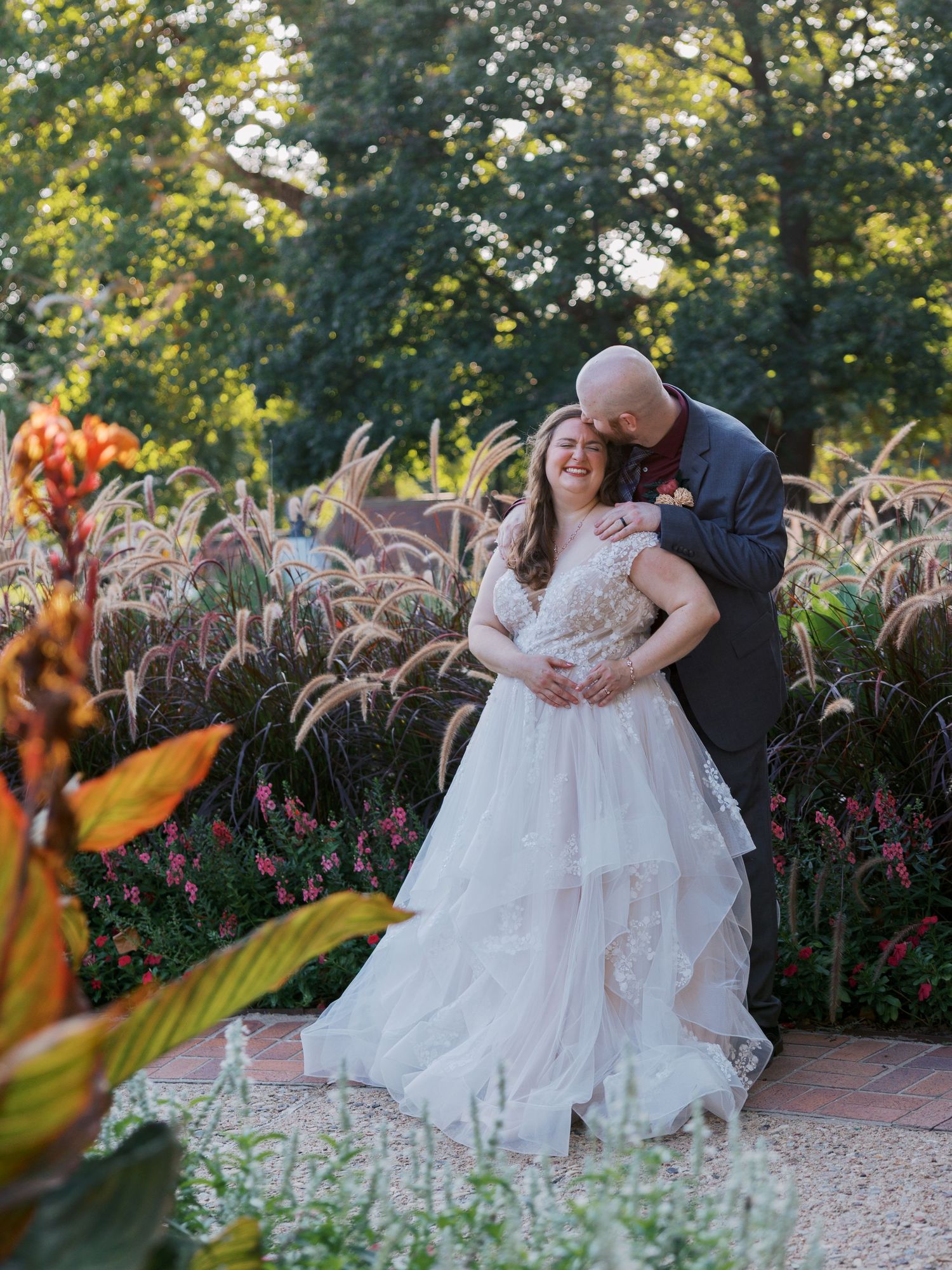 Bride in layered white gown embraced by groom in garden wedding portrait surrounded by colorful flowers and ornamental grasses.