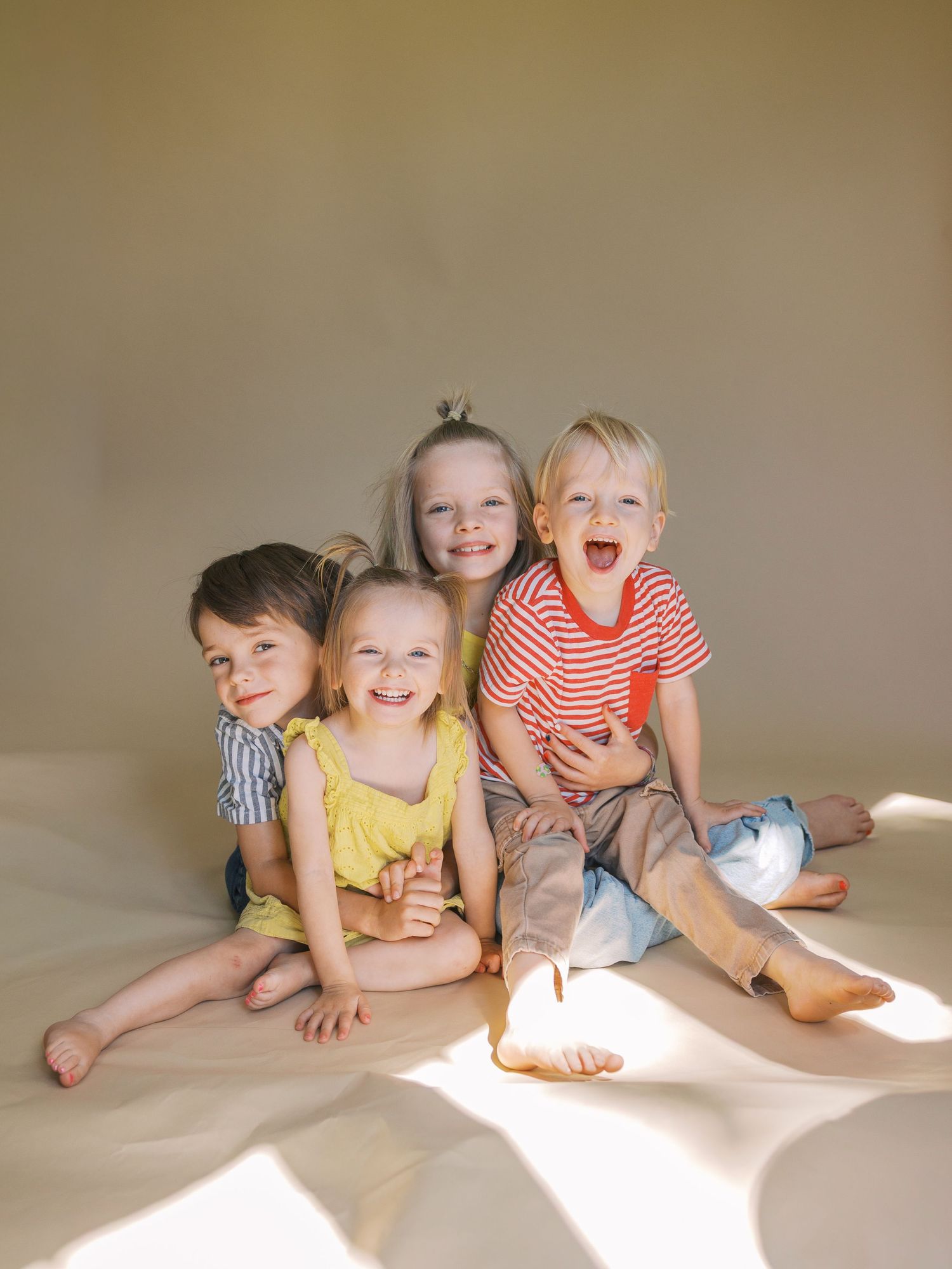 Four happy young children sitting together on a light floor, smiling and laughing in a bright, warmly lit portrait.