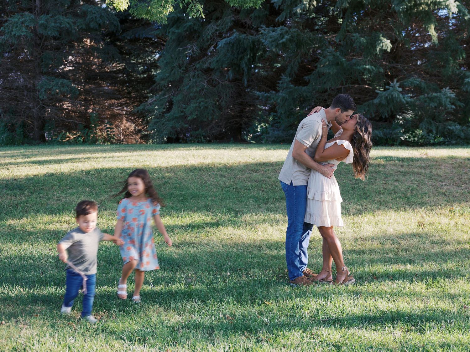 Family of four enjoying a sunny day in a lush green park, parents kissing while two young children play nearby.
