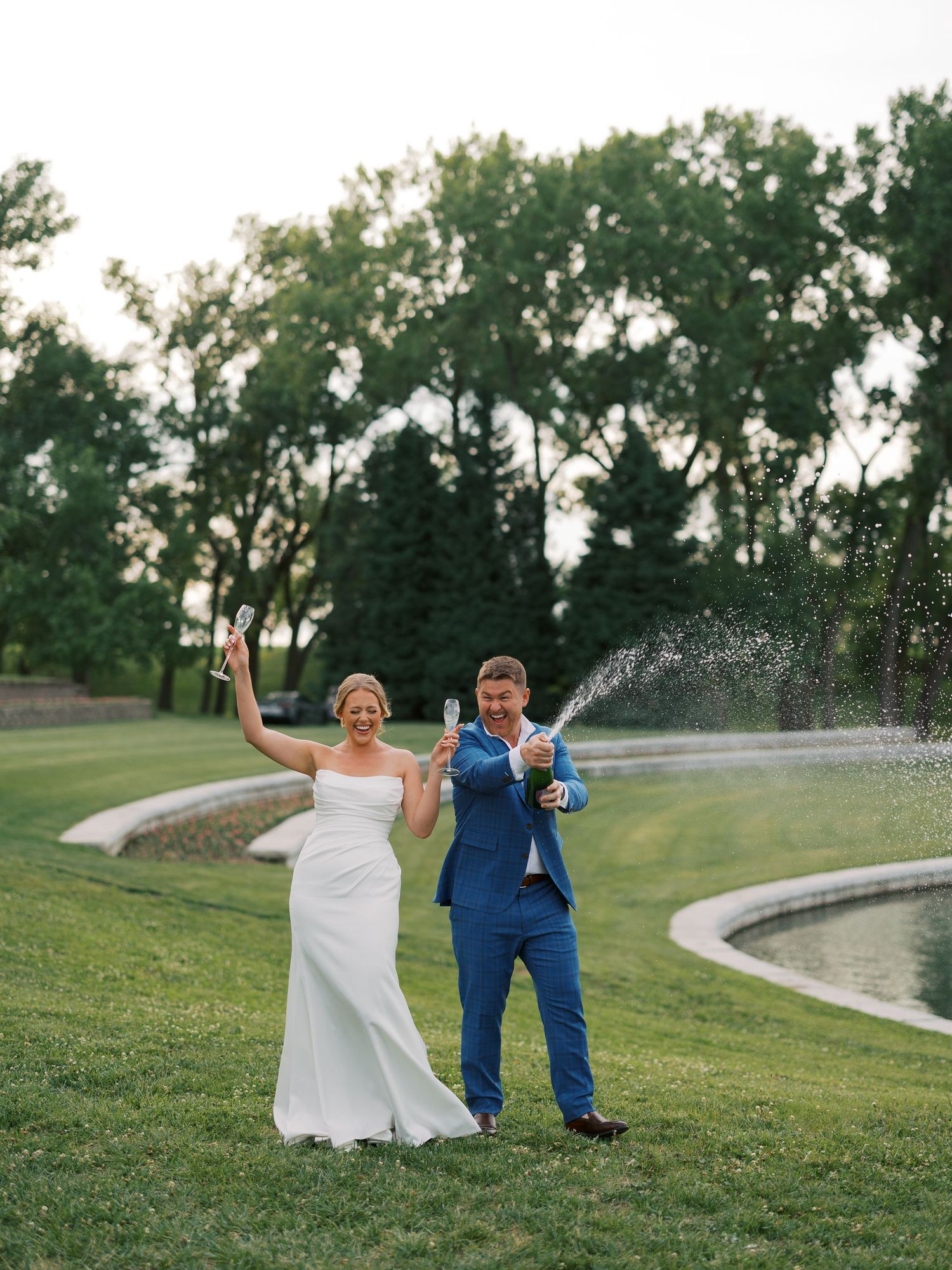 Bride and groom celebrating on golf course lawn, popping champagne bottles near a pond, surrounded by lush green trees.