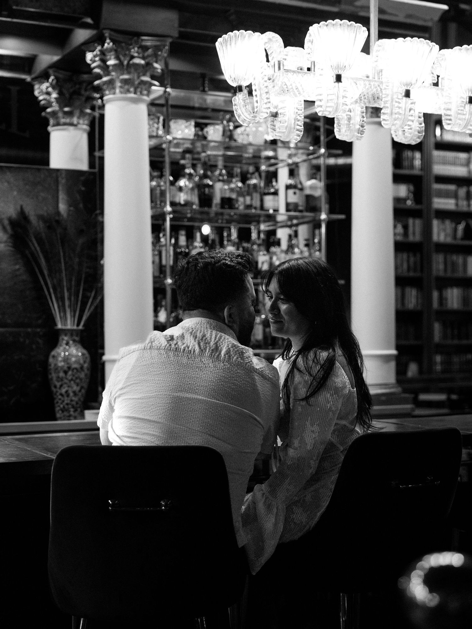 Black and white photo of a couple sharing an intimate moment at a bar with elegant chandeliers and shelves.