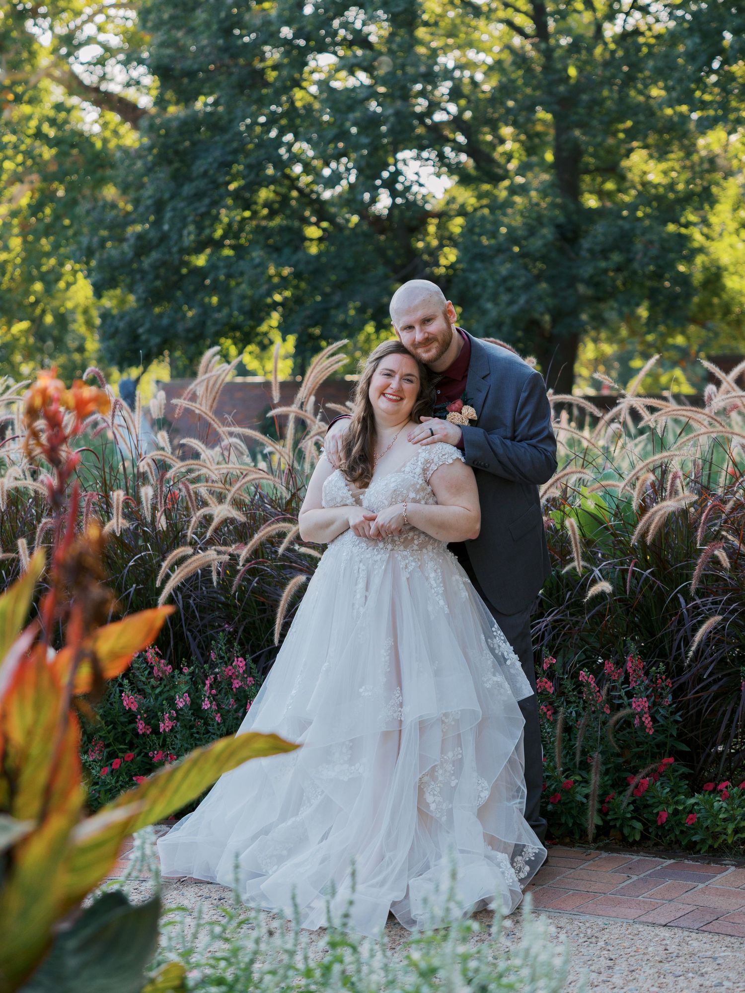A bride in a white lace gown and groom in a gray suit embrace in a lush garden with colorful fall flowers.