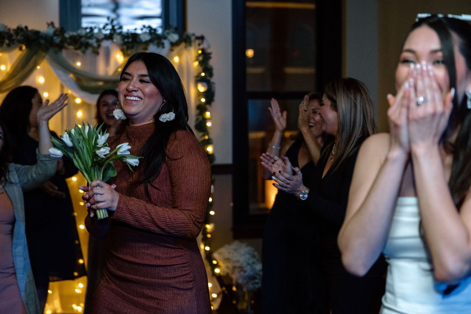 Women celebrating indoors with string lights, one holding white flowers, another covering her mouth in surprise.