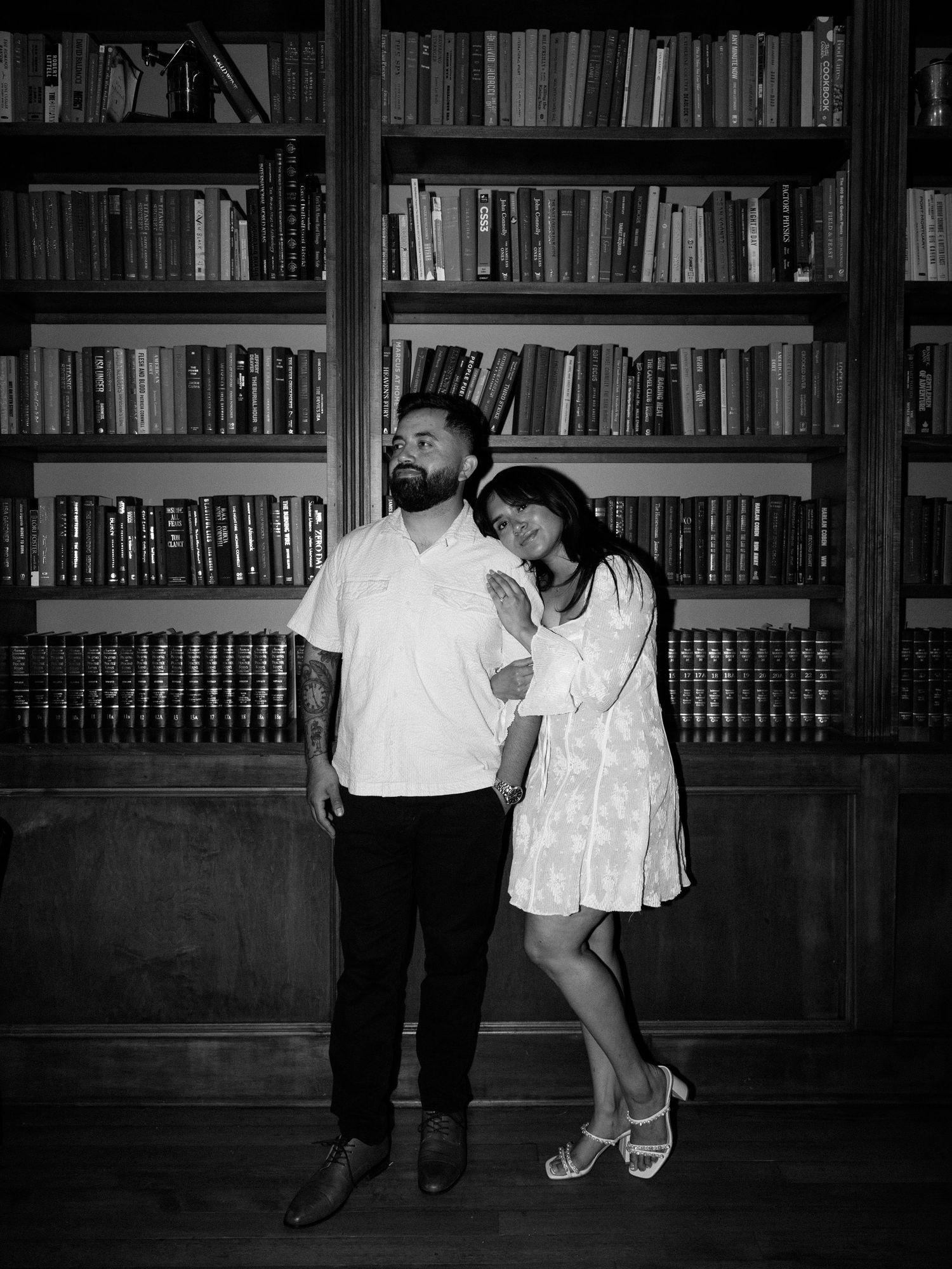 A couple poses playfully against wooden bookshelves in a library setting.