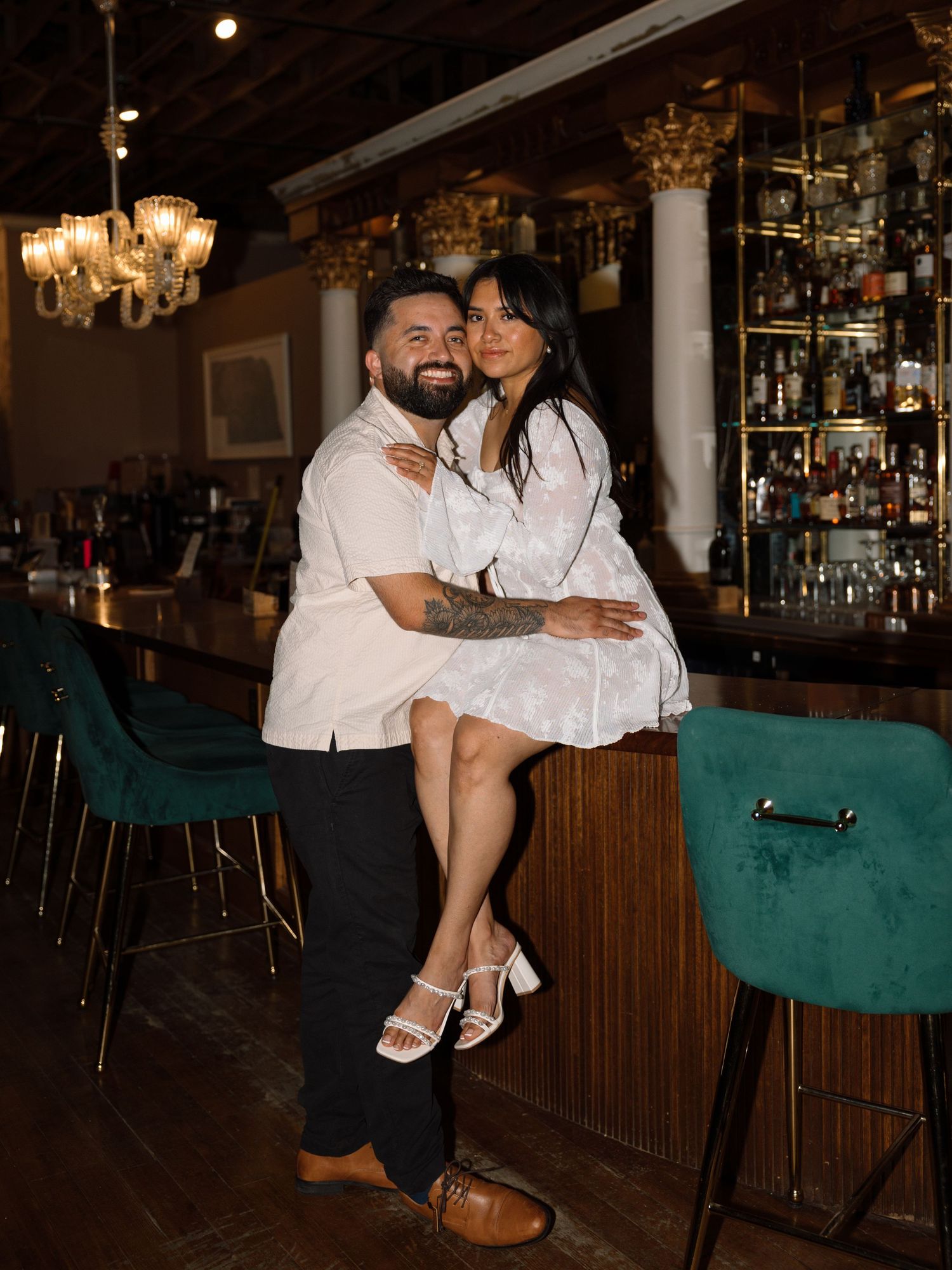 Couple embracing at an elegant bar with green velvet stools, bottle-lined shelves, and chandelier lighting.