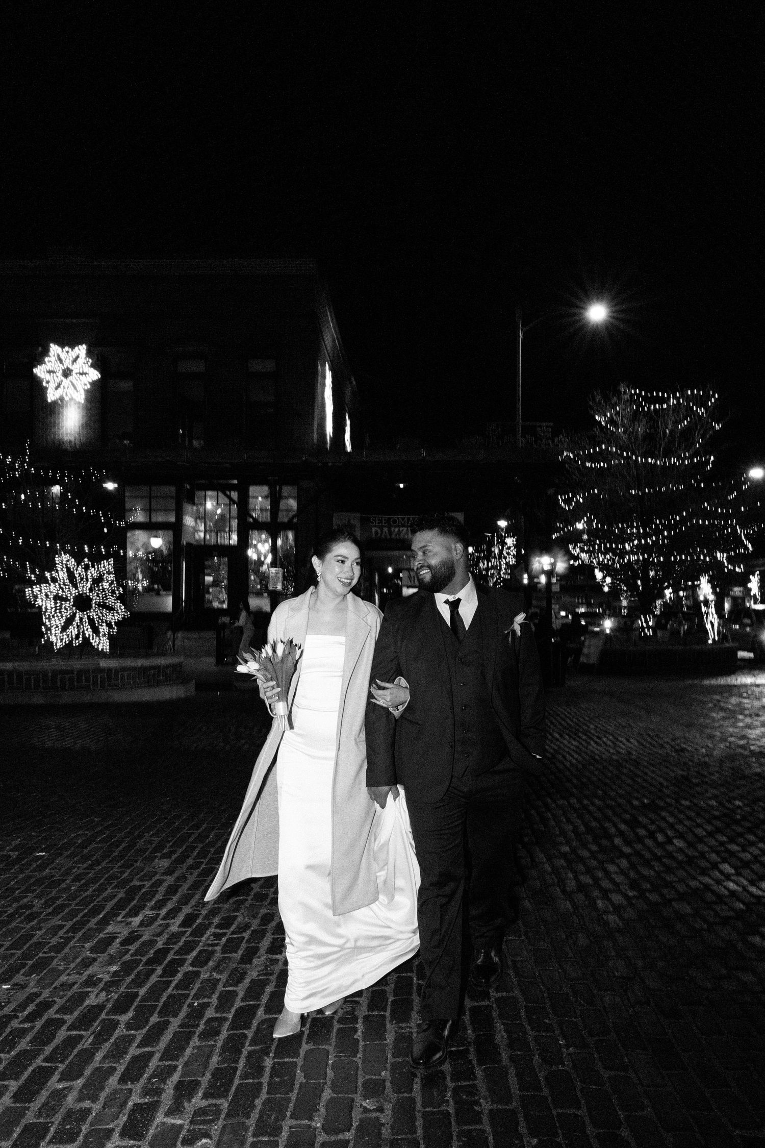 Black and white nighttime photos of a couple walking on cobblestone streets with decorative lights in the background.