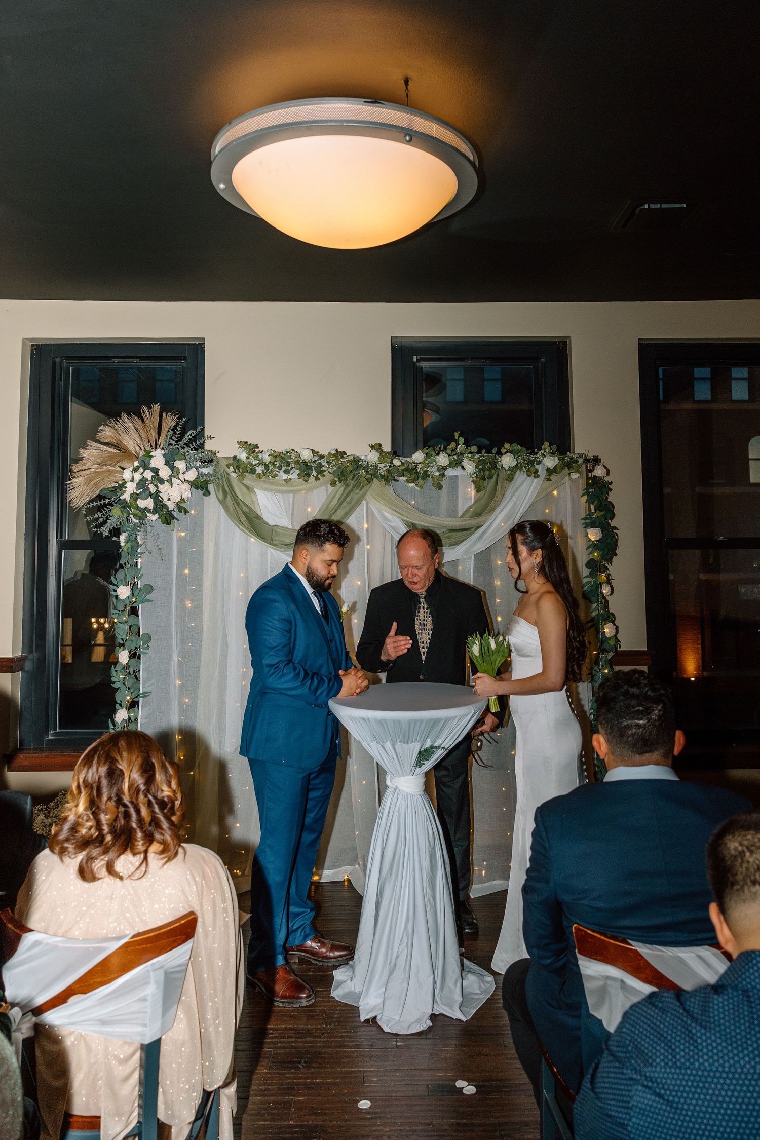 Couple exchanging vows at indoor wedding ceremony with floral arch, officiant, and seated guests.