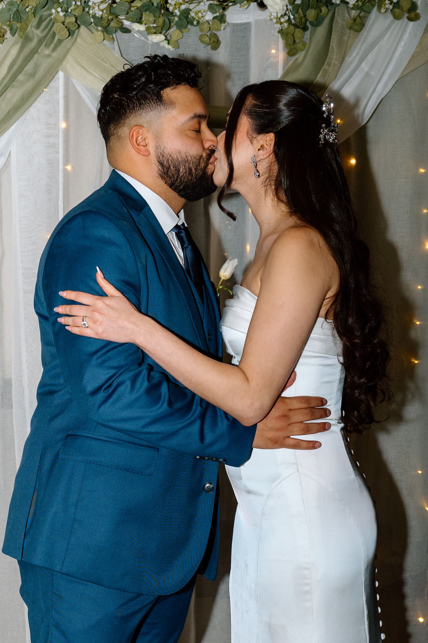 Bride and groom share first kiss at wedding altar, groom in blue suit, bride in white gown with floral backdrop.