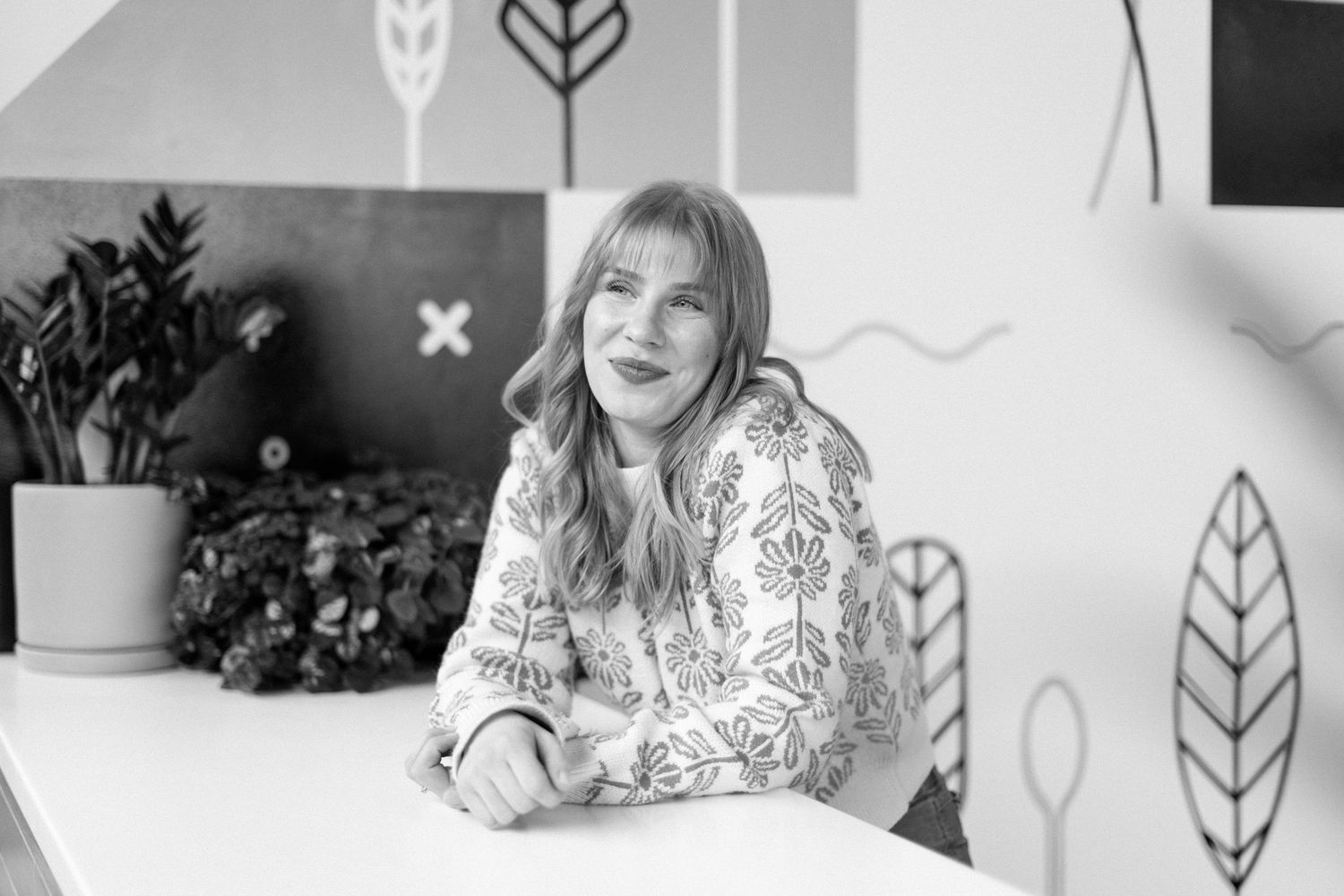 Black and white portrait of a smiling woman with long hair sitting at a white desk in a modern creative office.