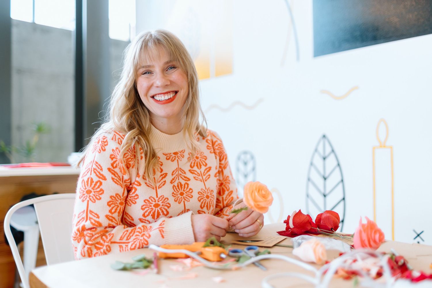 Smiling blonde woman in orange floral top crafting with flowers at a white table in a bright studio.