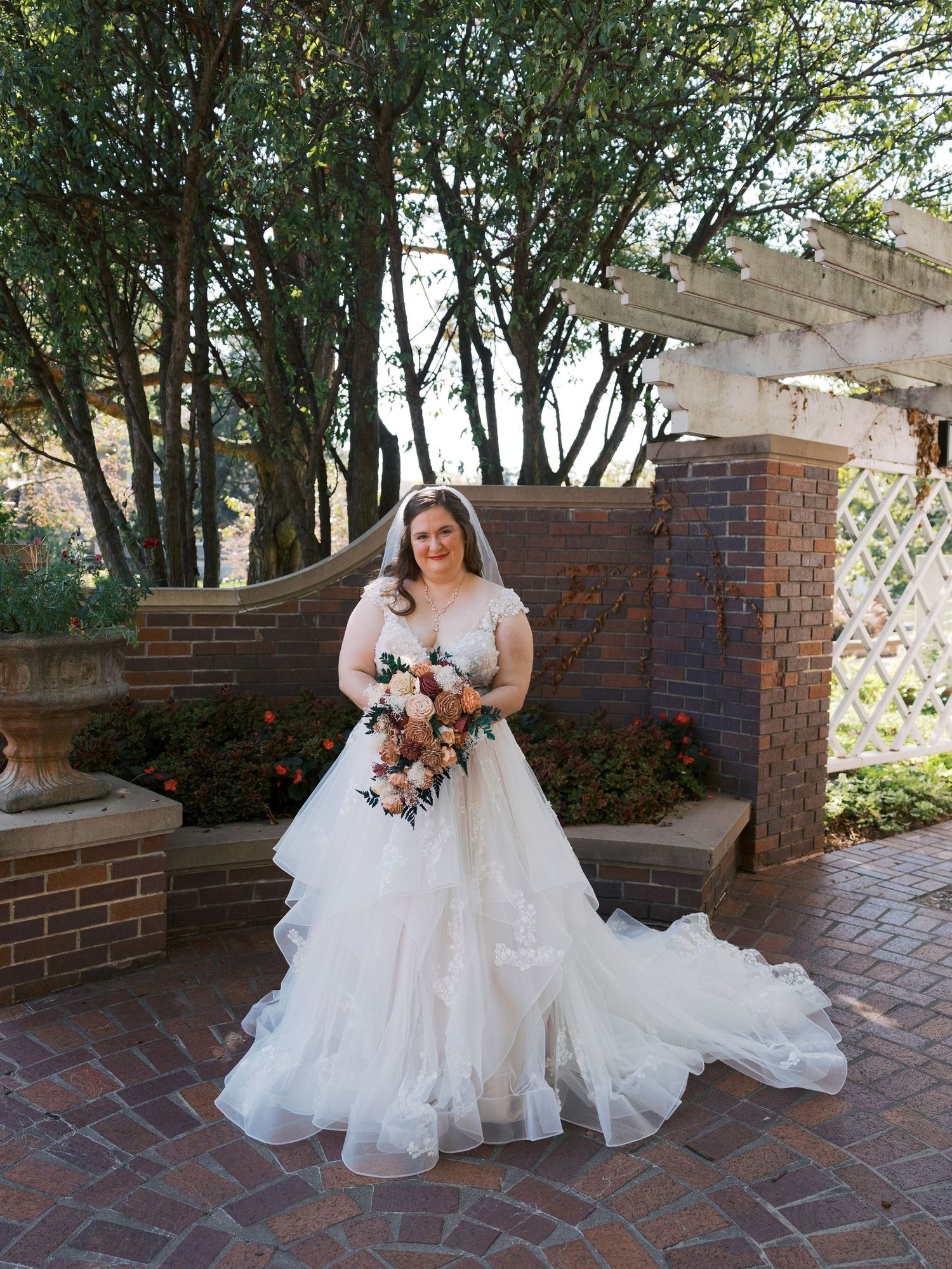 Bride in a white ball gown with tulle skirt holds floral bouquet, standing on brick patio with garden arbor backdrop.