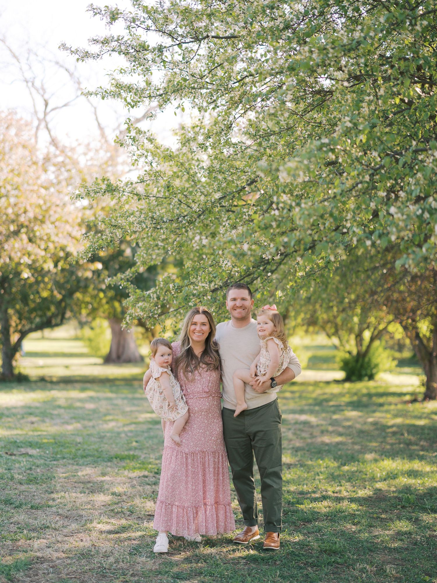 Family of four posing under blooming spring trees in a sunlit park, parents holding young children in coordinated outfits.