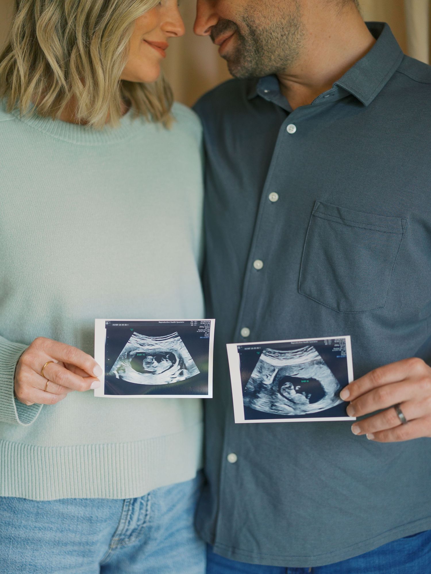 Couple holding ultrasound photos together, announcing pregnancy with matching teal outfits in a soft, natural light setting.