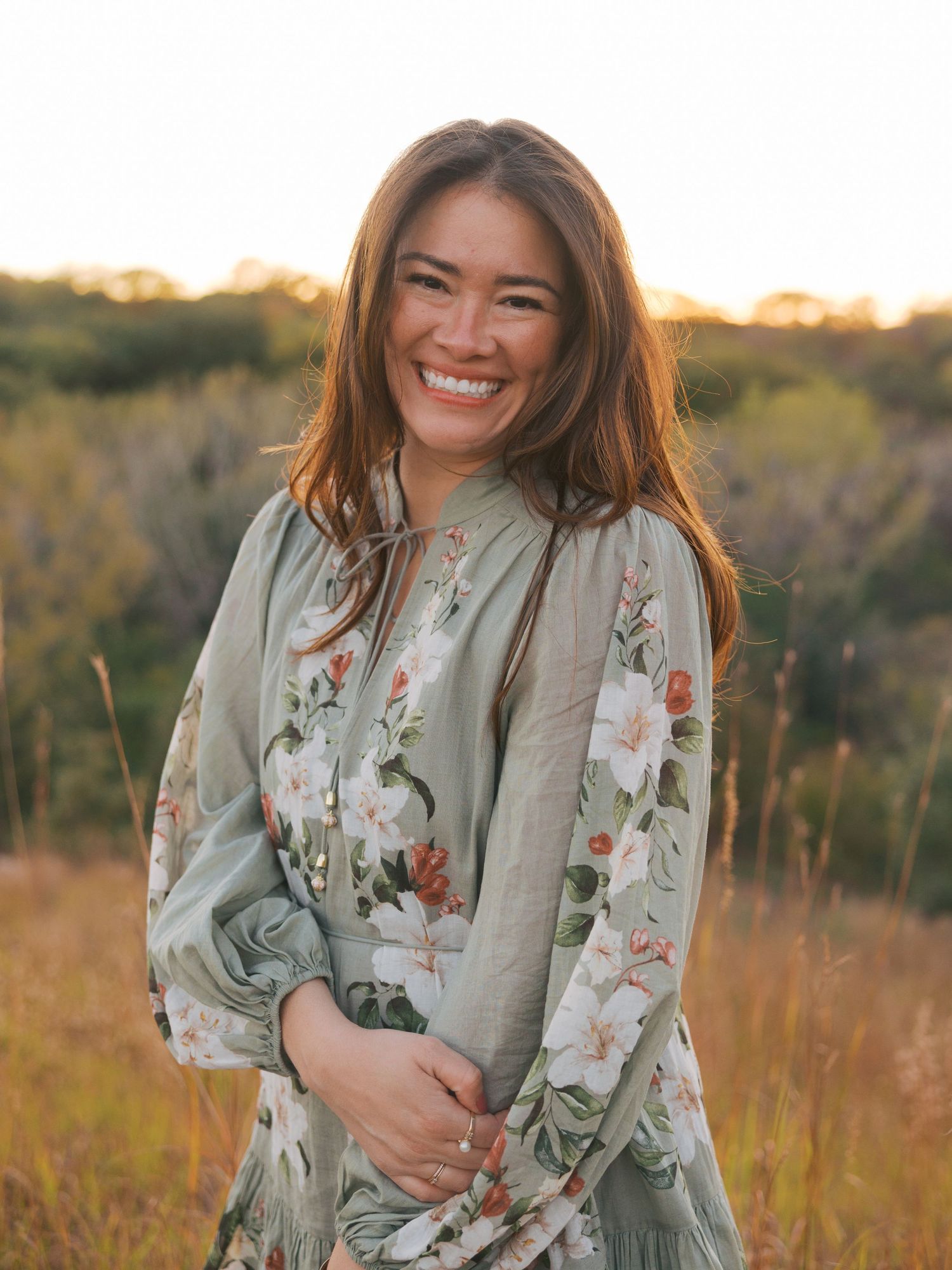 Smiling woman in floral sage green dress standing in golden field at sunset.