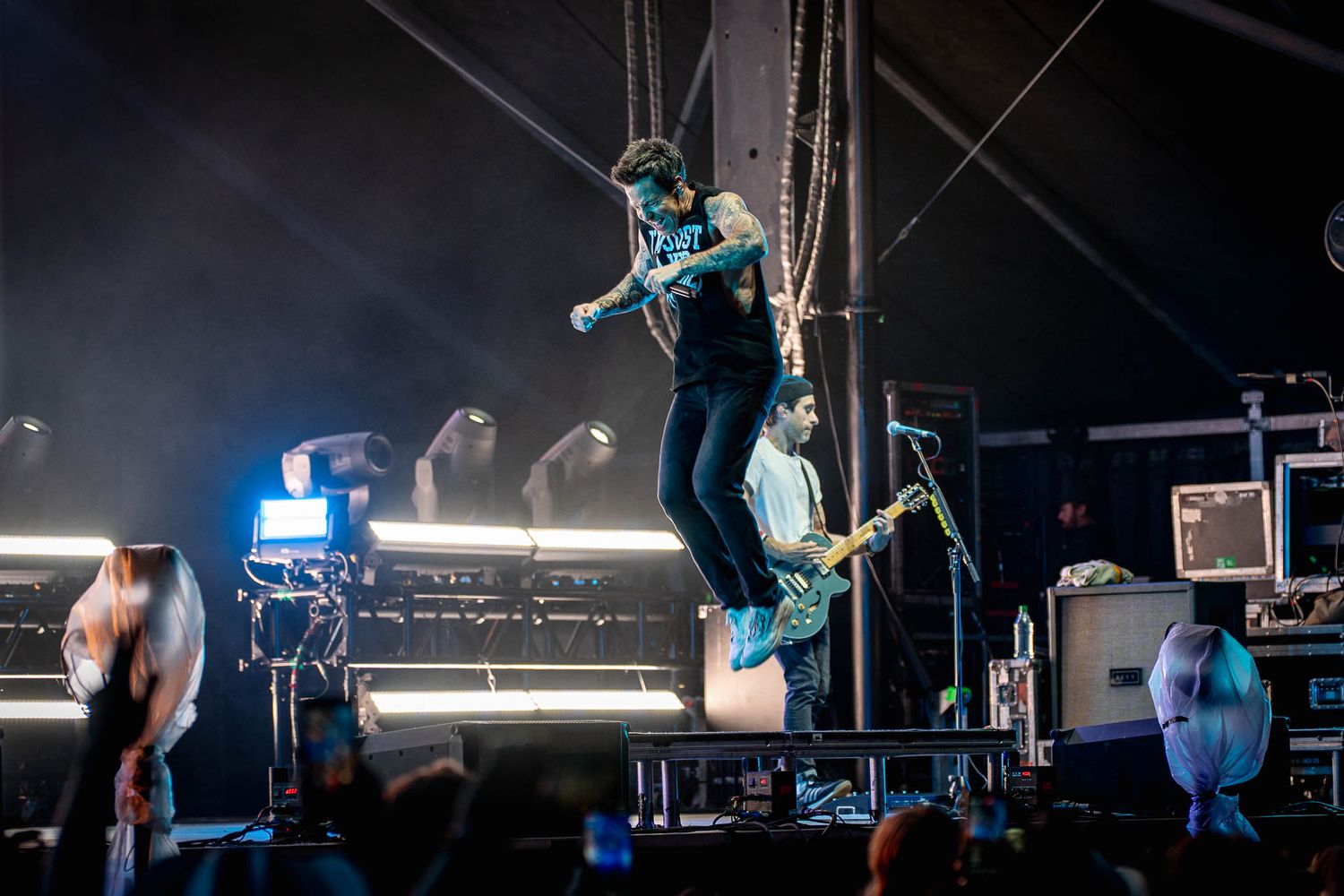 Simple Plan in blue outfit leaps high during energetic concert performance with stage equipment visible.