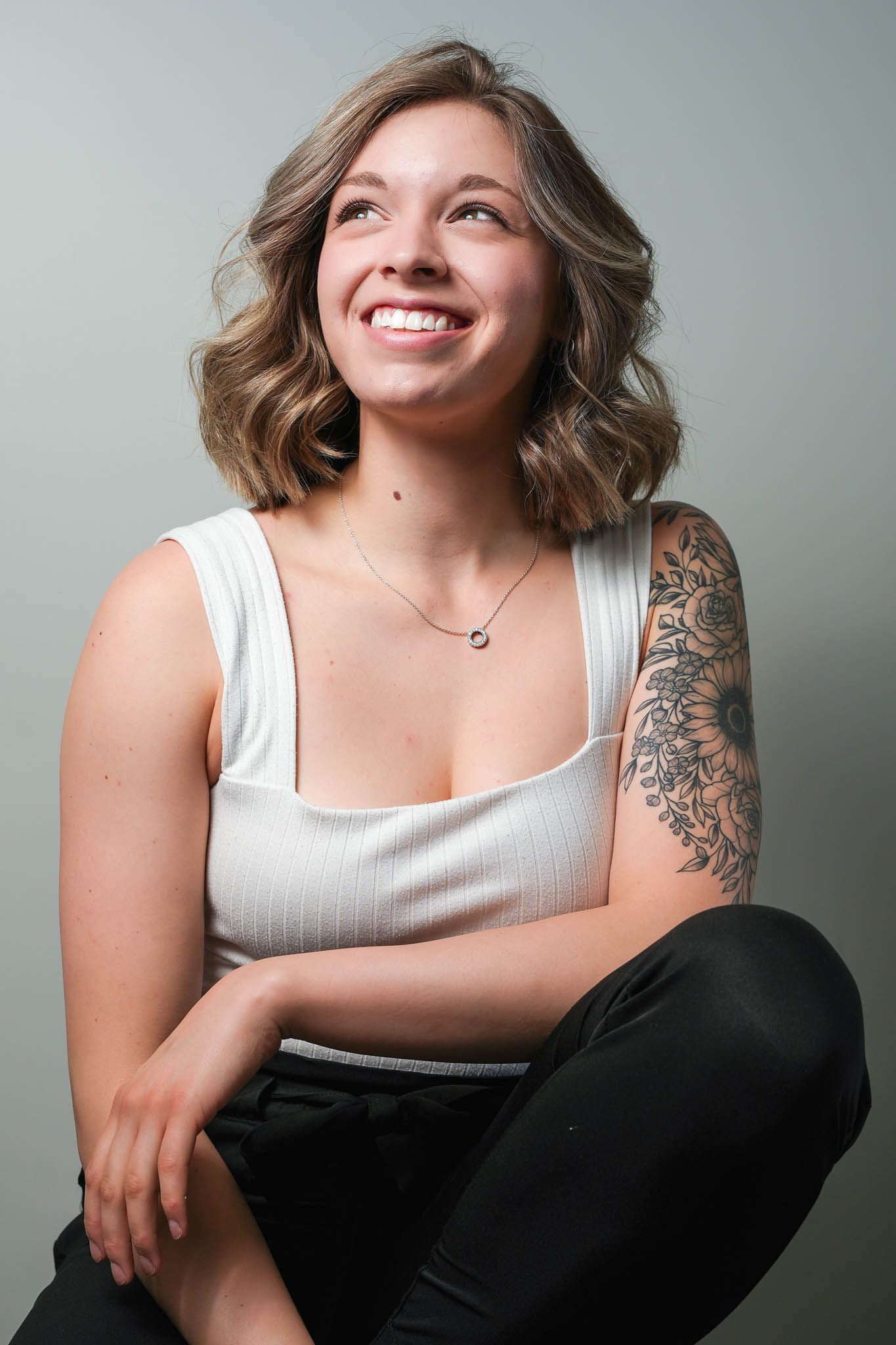 Person in white tank top with shoulder tattoo poses casually against grey wall.