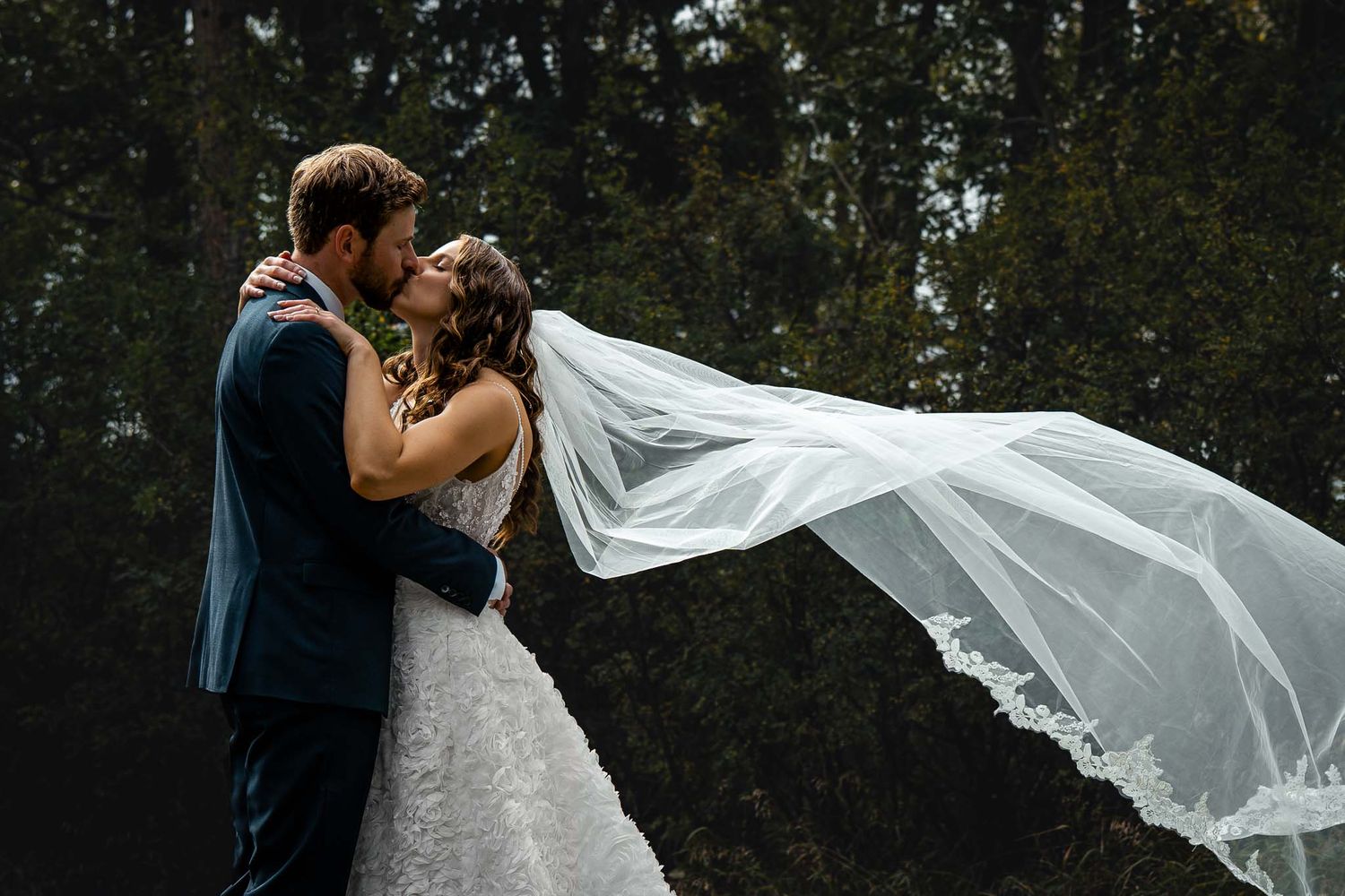 Wedding couple poses dramatically with flowing veil in natural setting.