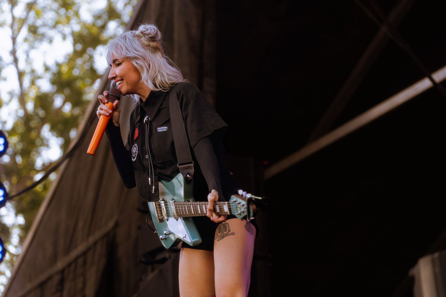 Stage performer in dark outfit plays guitar during outdoor daytime concert.