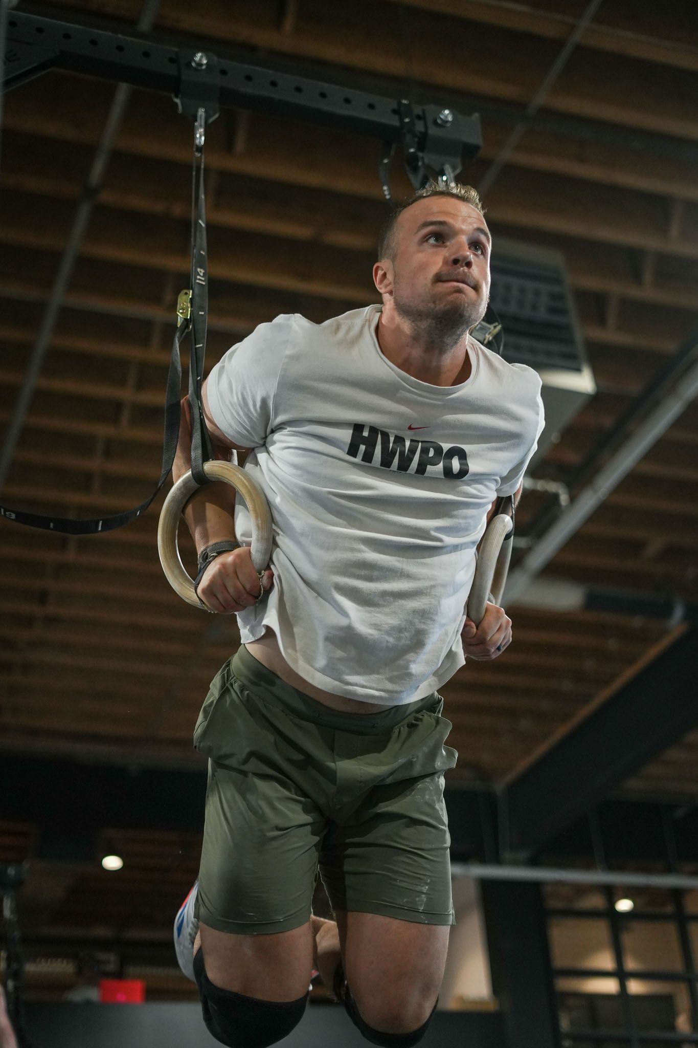 Athlete performing gymnastic rings exercise sequence in gym wearing white t-shirt and olive shorts.