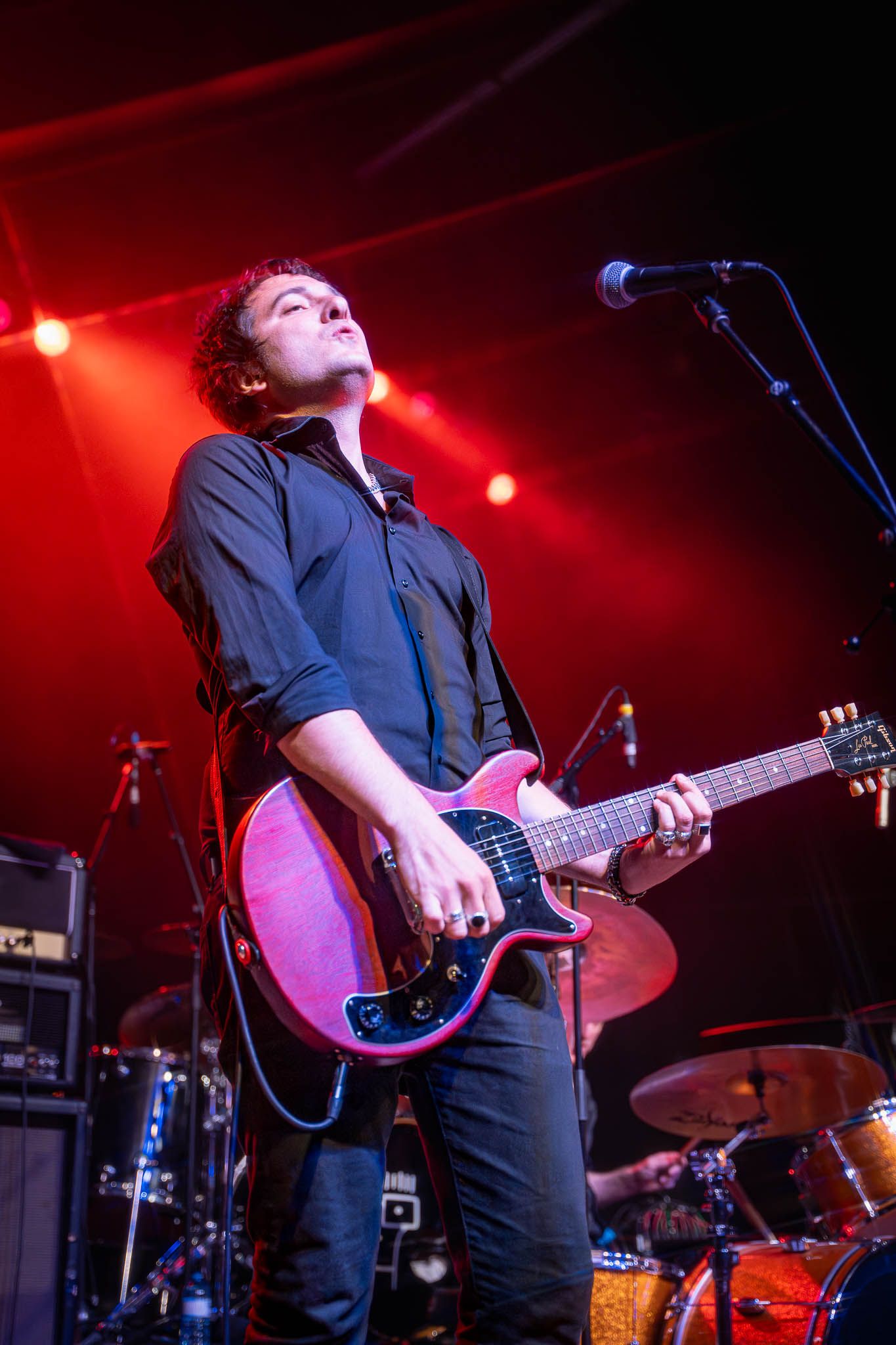Guitarist performing under red stage lighting with musical equipment visible.