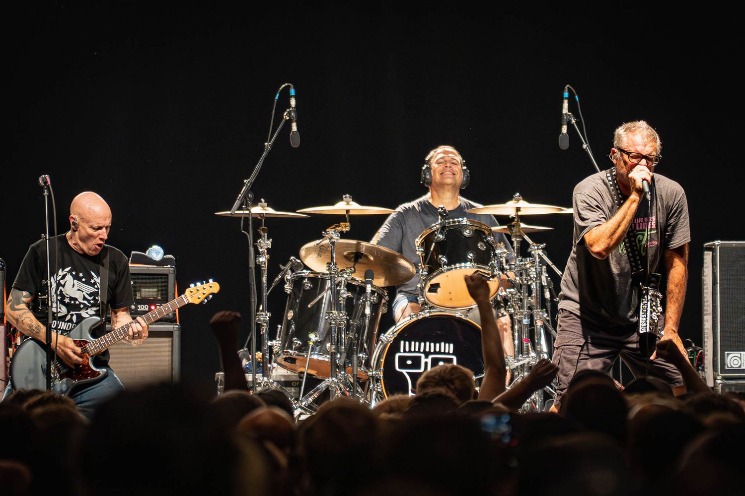 Panoramic view of The Descendents band performing on dimly lit stage with instruments and equipment.
