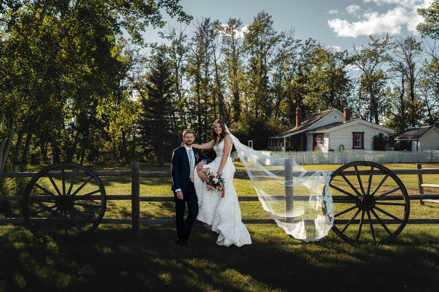 Romantic wedding couple poses in a rustic outdoor setting with wagon wheels and white buildings in the background.