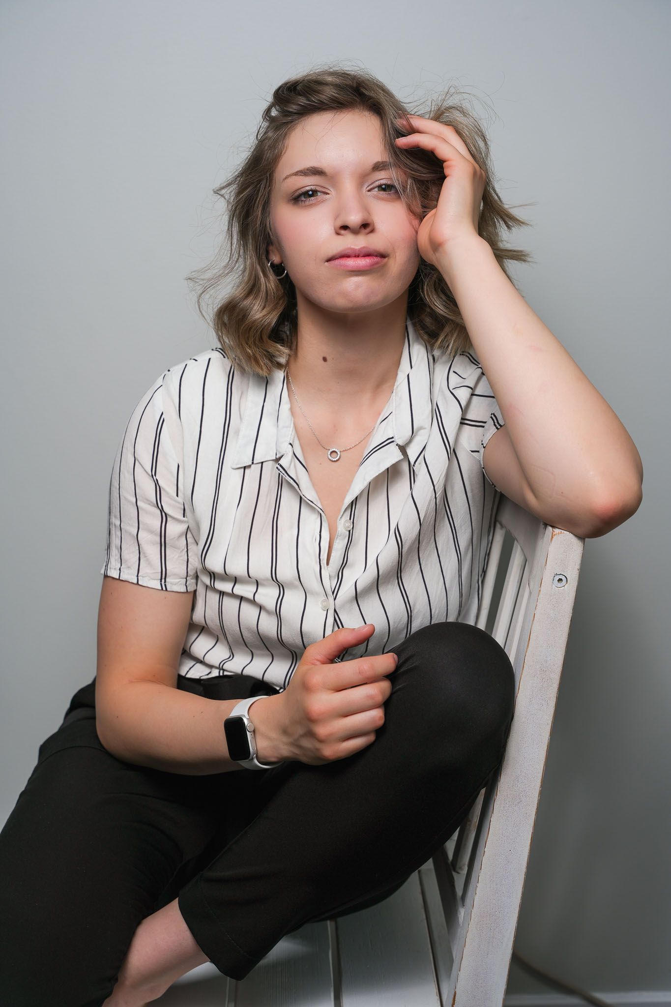 Person in striped shirt poses casually on chair against grey backdrop.