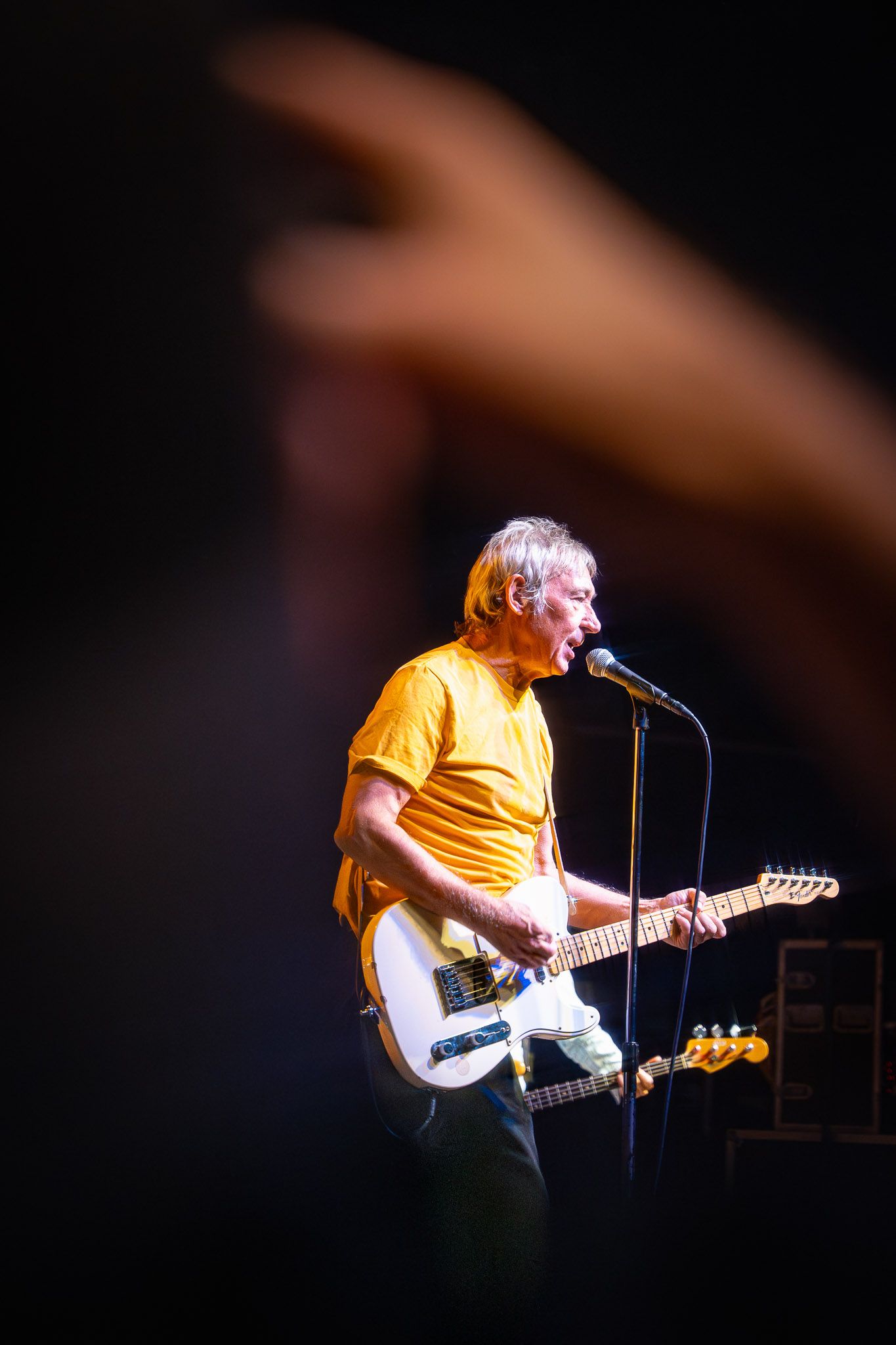 Guitarist from The Buzzcocks in yellow t-shirt performs energetically on stage with electric guitar under dramatic lighting.
