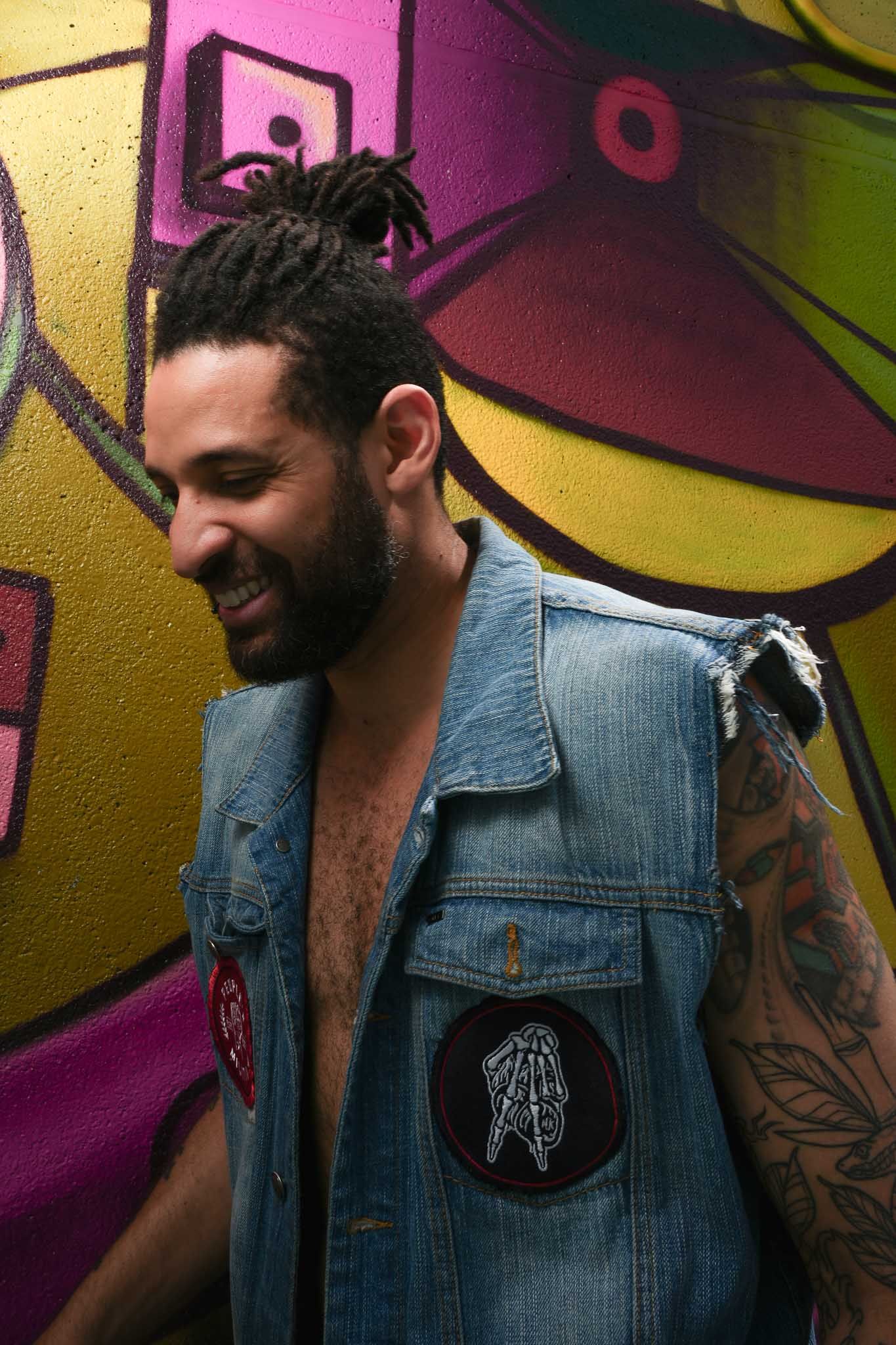 Tattooed person in denim vest poses against colorful graffiti wall with artistic lighting.