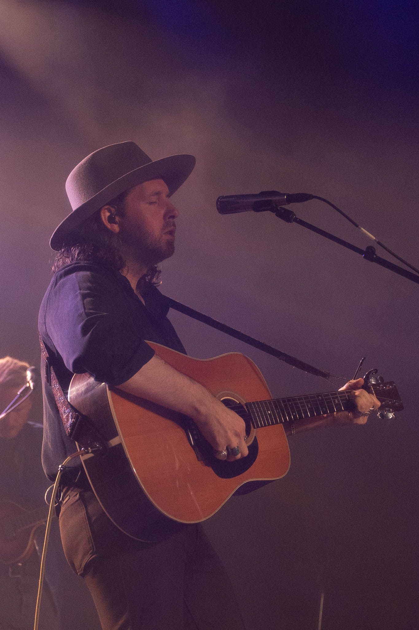 Musician in cowboy hat playing acoustic guitar while singing on dimly lit stage.
