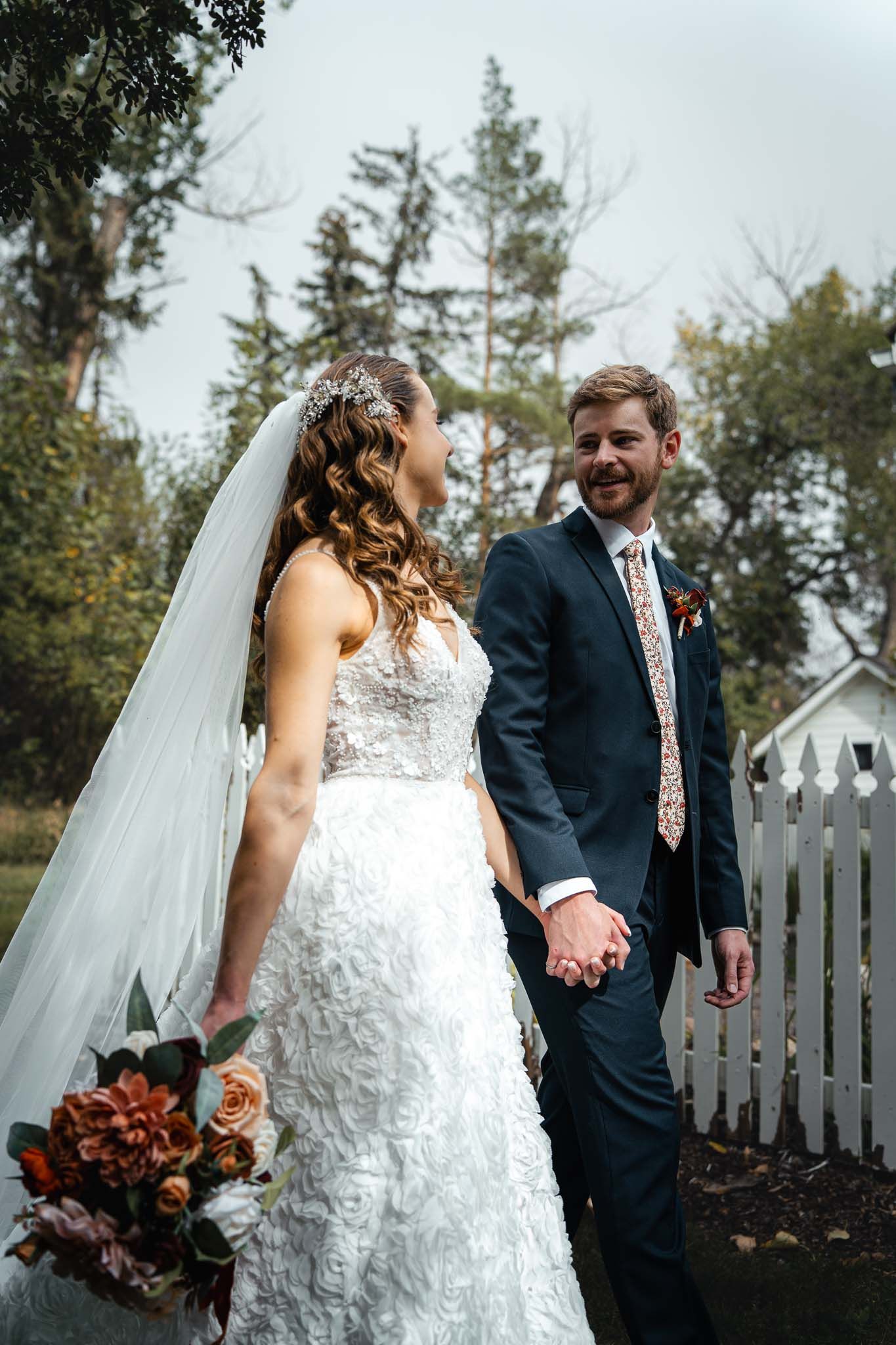 Newlyweds share candid moments walking along white picket fence.