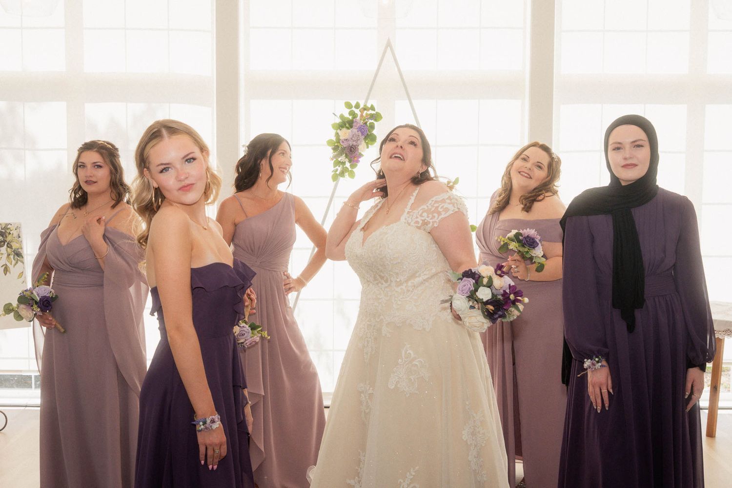 Wedding party poses together in coordinated purple and mauve dresses by a bright window.