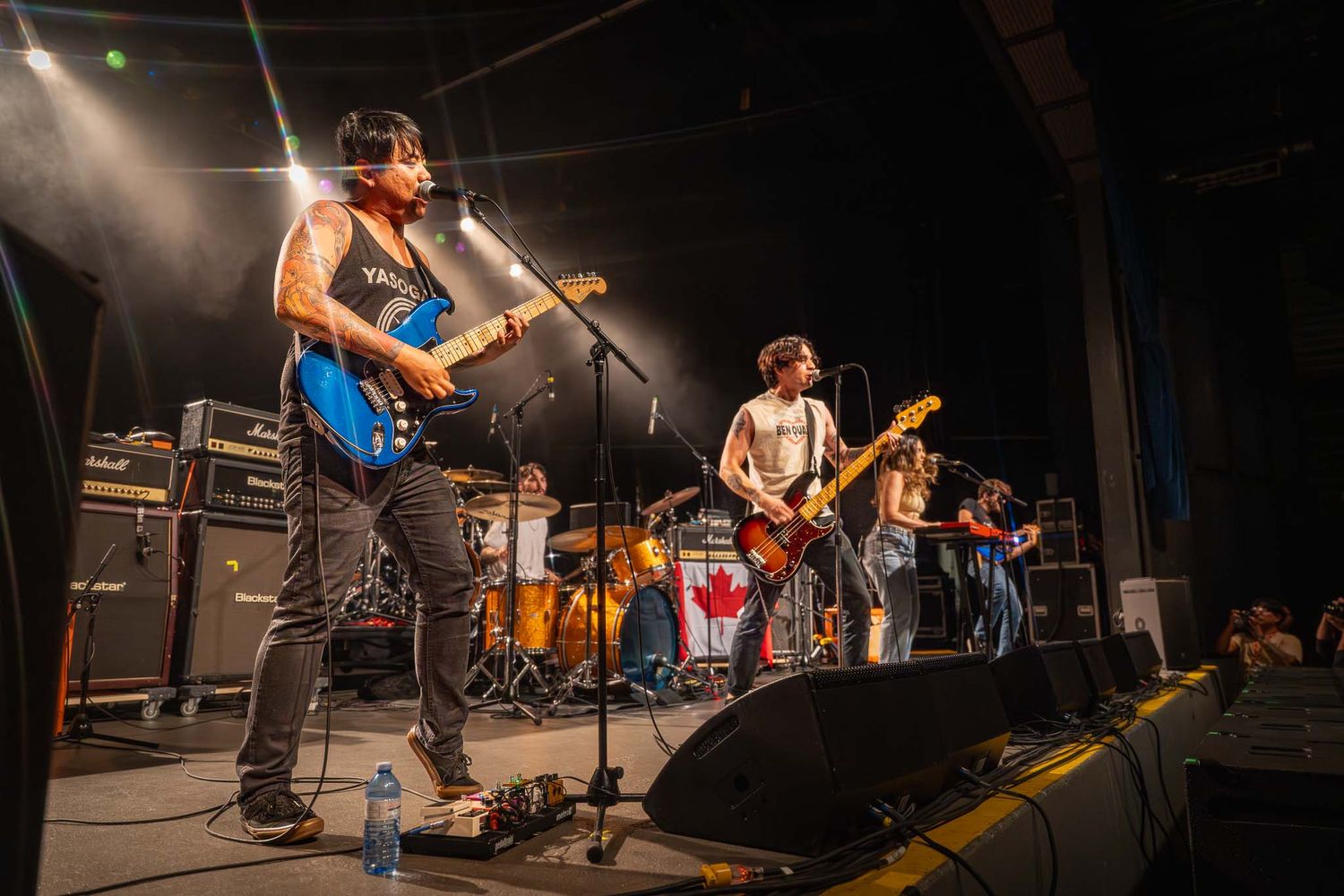 Wide shot of band performing on dimly lit stage with instruments and equipment visible.
