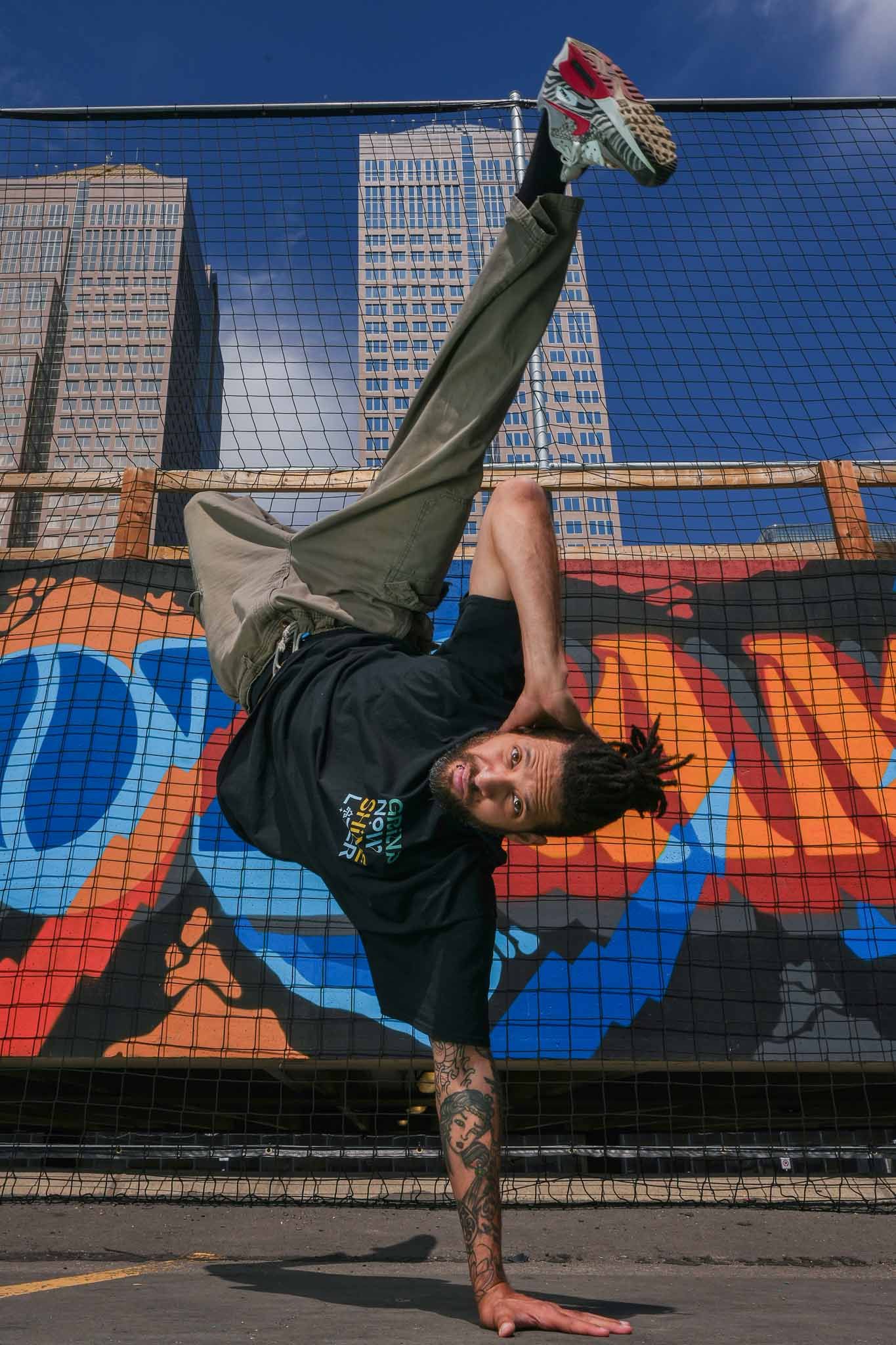 Breakdancer performs horizontal hold on railing with graffiti wall and buildings in background.