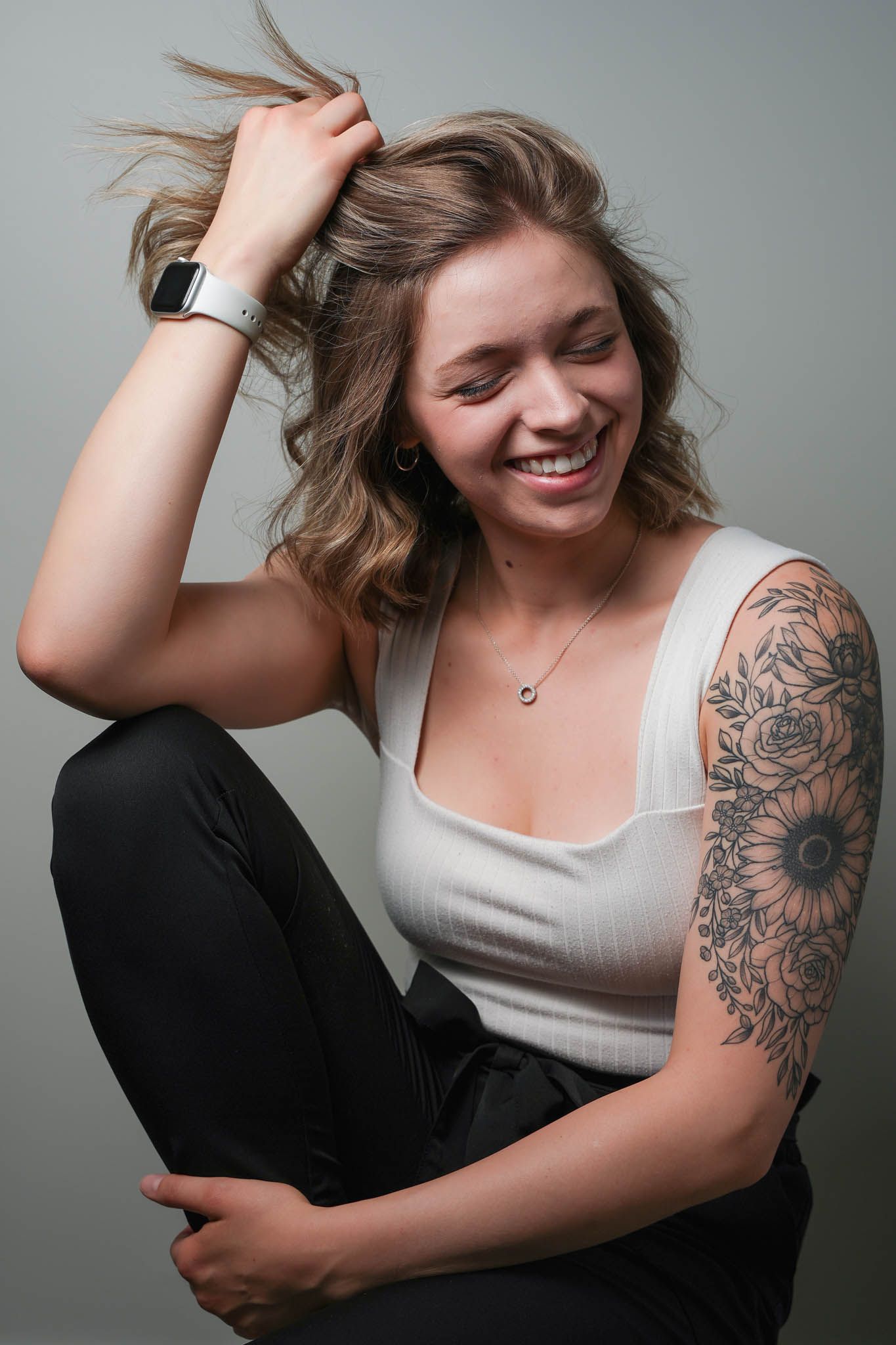 Person in white tank top with floral arm tattoo poses playfully against grey background.
