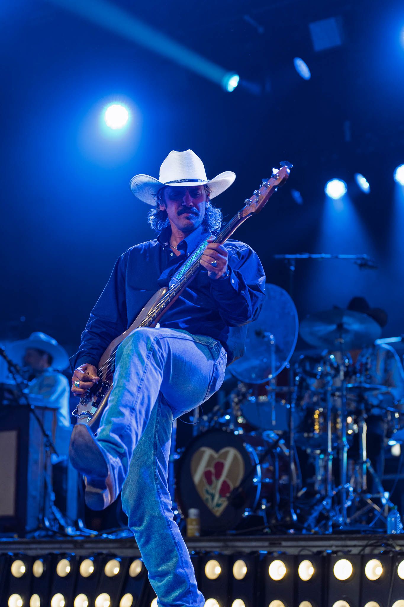 Performer in cowboy hat plays guitar on stage bathed in blue concert lighting.