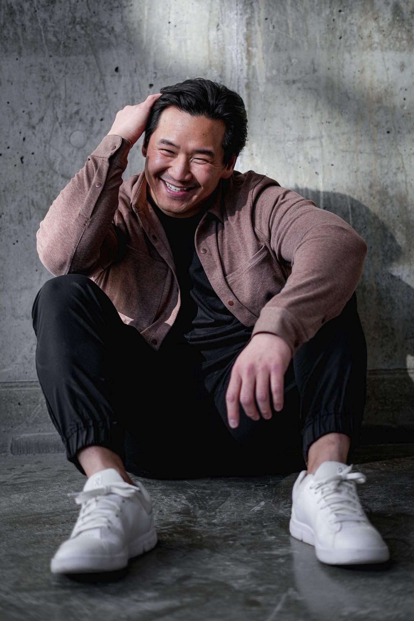 Casual portrait series showing person in brown sweater and black pants sitting on concrete floor with white sneakers.