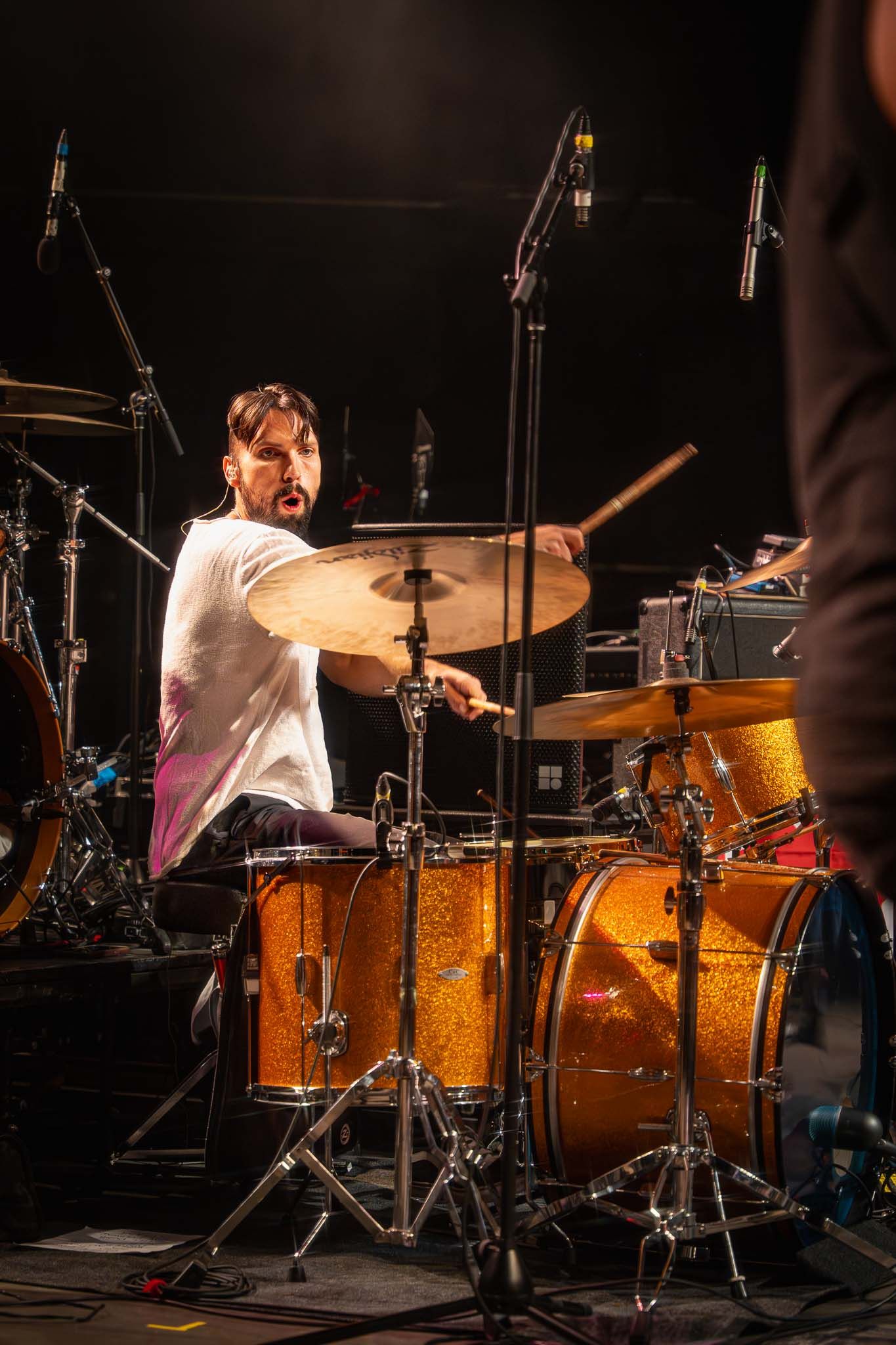 Drummer plays golden drum kit on dimly lit concert stage with dramatic spotlights.