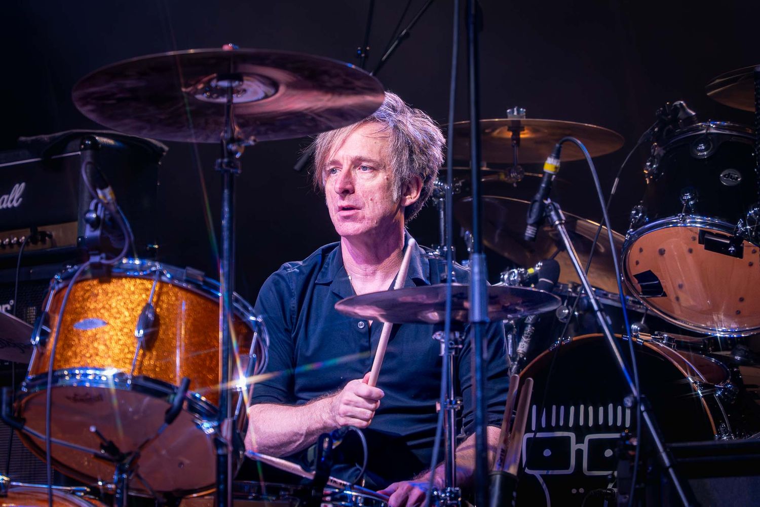 Drummer from The Buzzcocks performs behind a large drum kit on stage with colorful stage lighting in background.