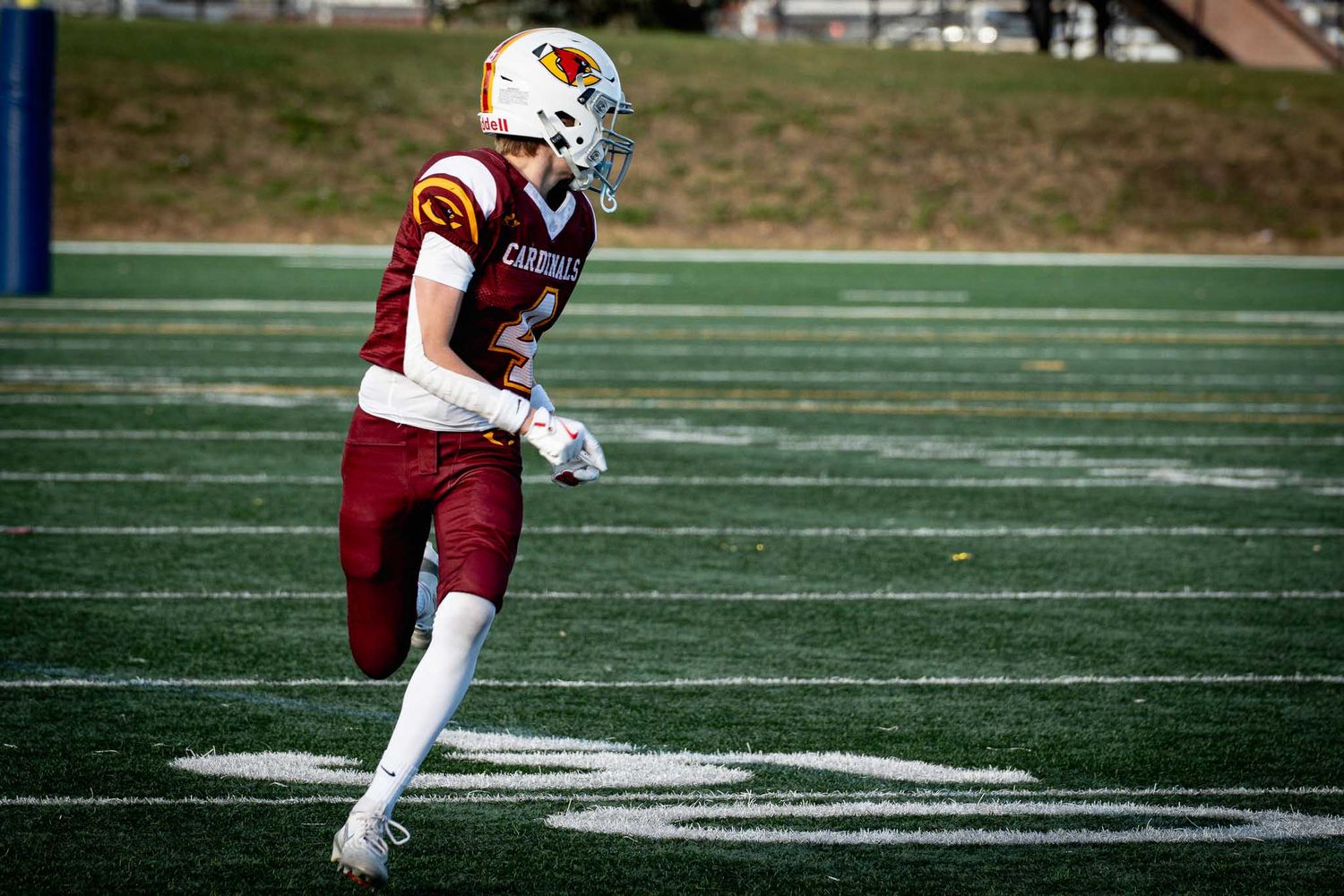 Football players in white and maroon uniforms running plays on artificial turf field.