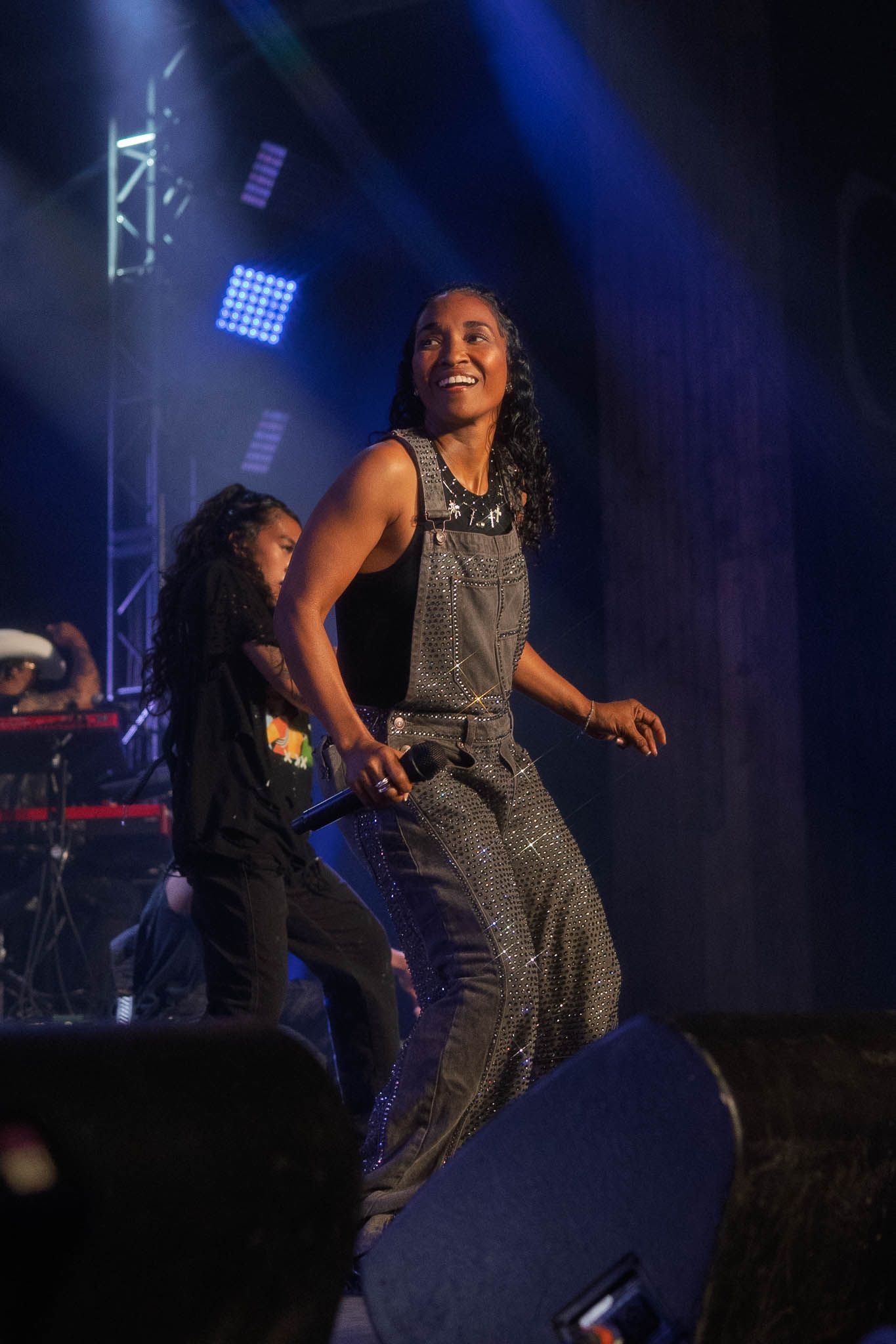 Performer in sparkly overalls dances energetically on dark stage with dramatic lighting.