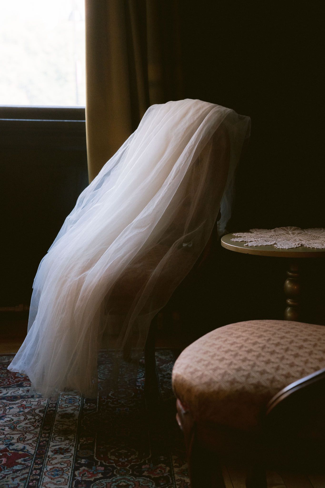 Elegant white wedding dress with tulle skirt draped over an upholstered chair in a dimly lit room.