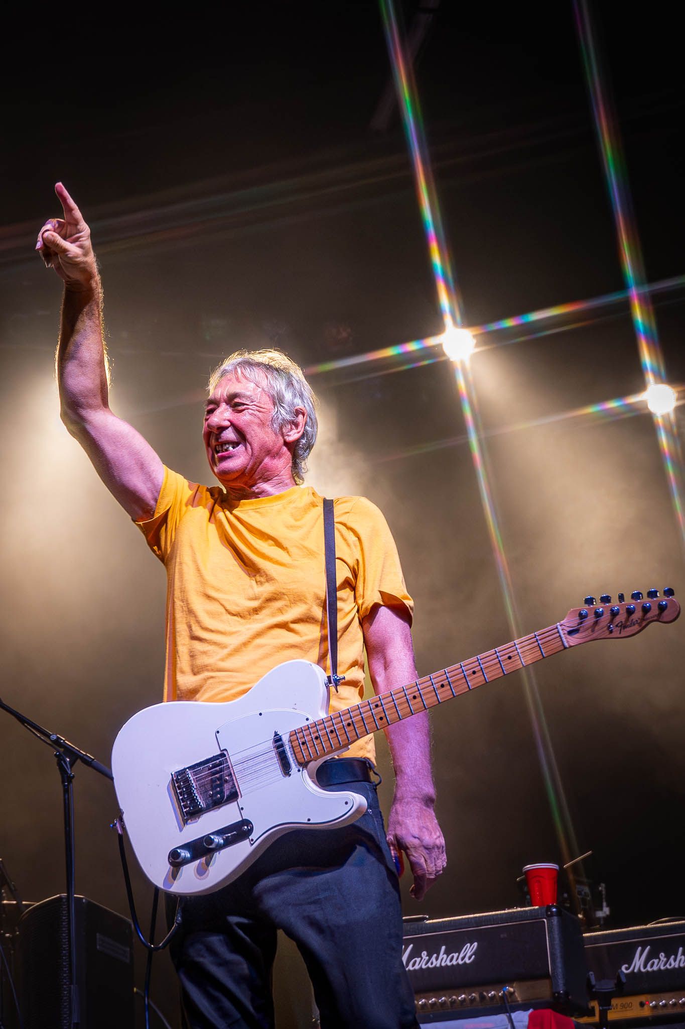 Performer in yellow shirt plays white electric guitar while singing on concert stage.