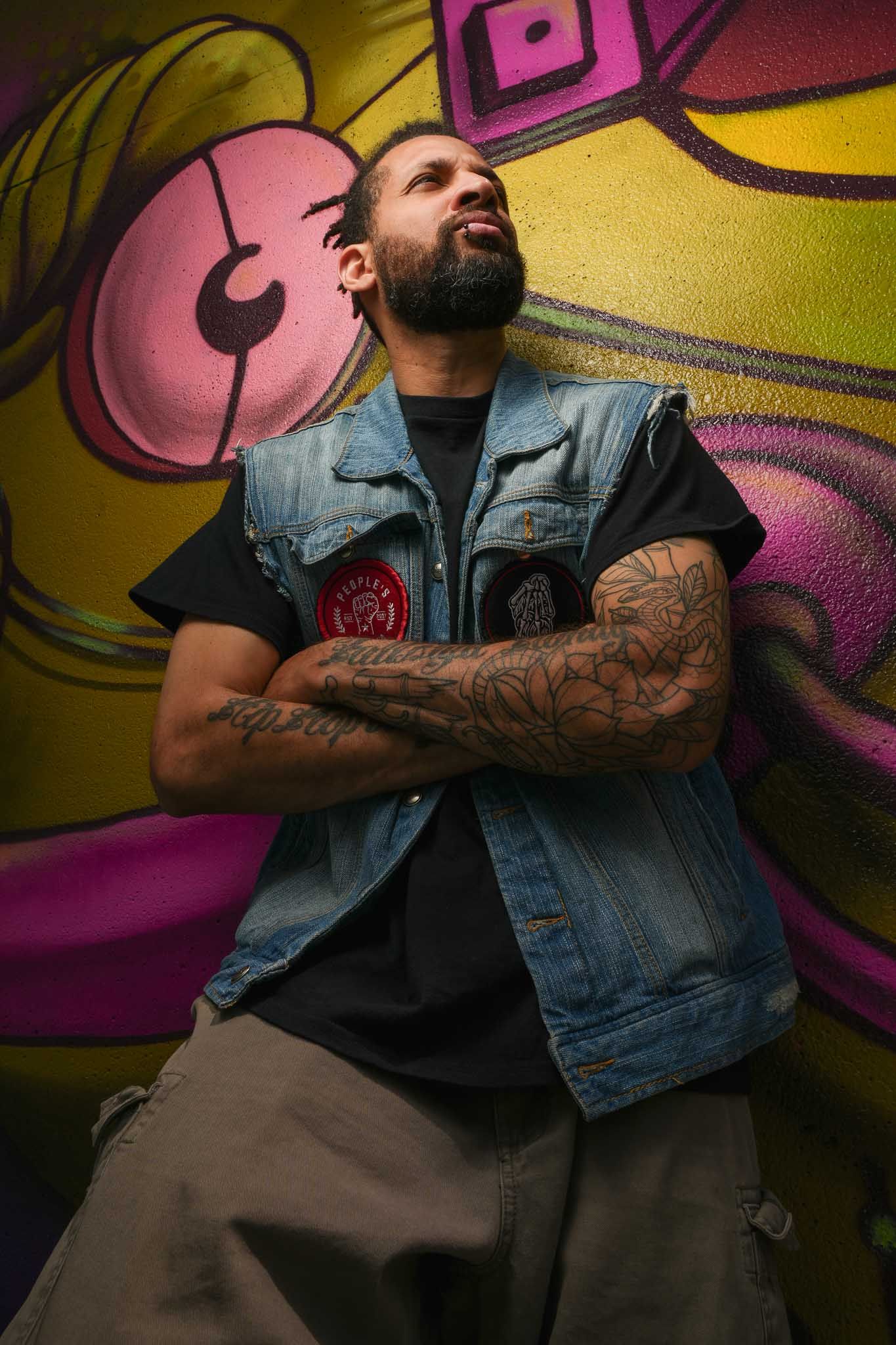 Dancer strikes expressive pose against vibrant graffiti wall wearing denim vest.