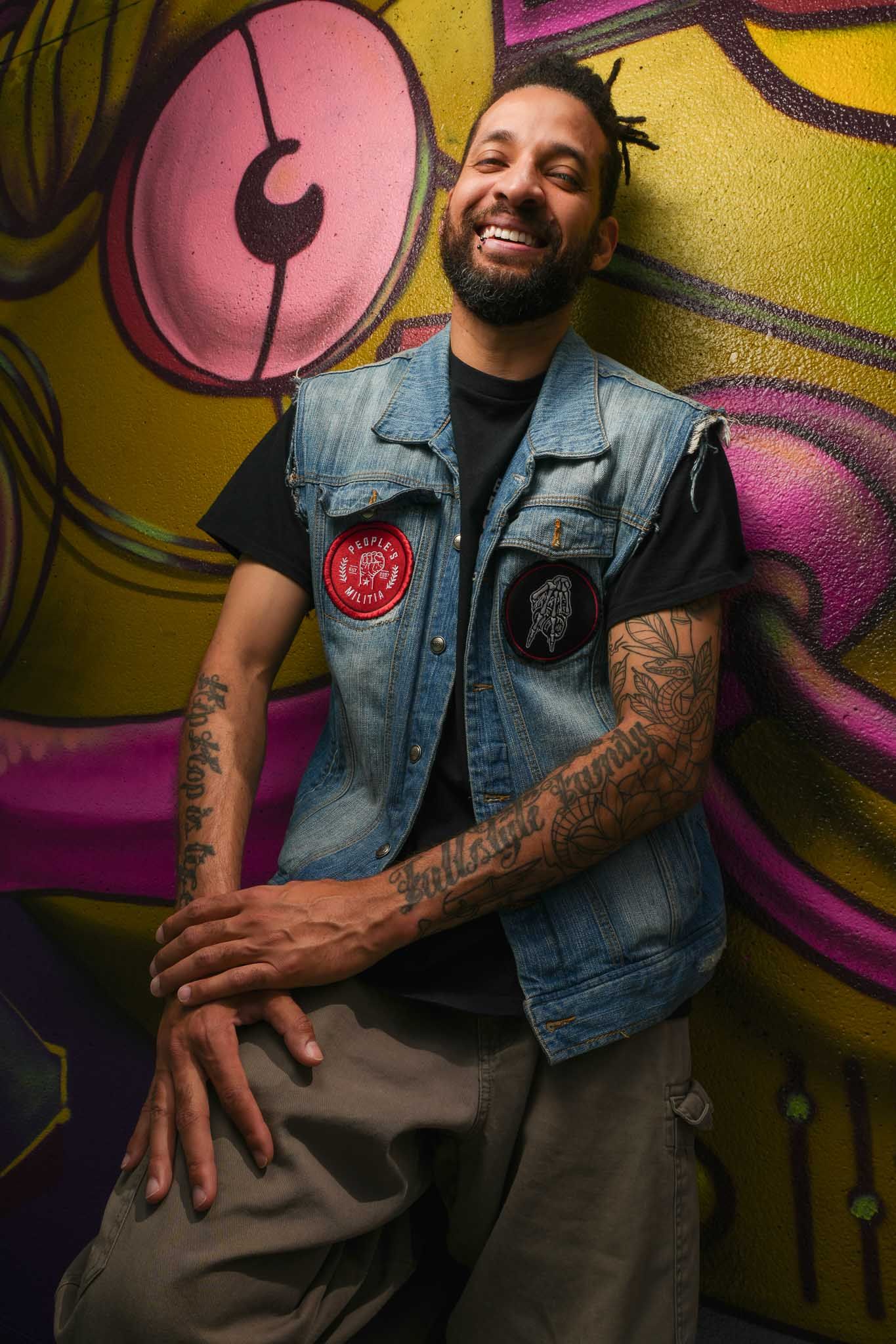 Performer poses expressively against yellow and pink graffiti wall wearing denim vest.