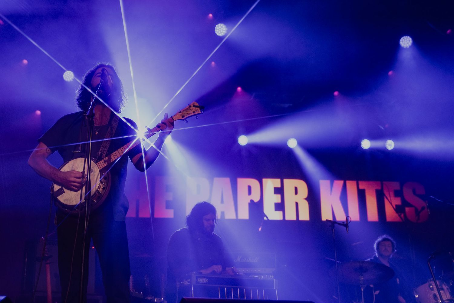 Concert stage bathed in deep blue lighting with band name 'Paper Kites' visible in background.