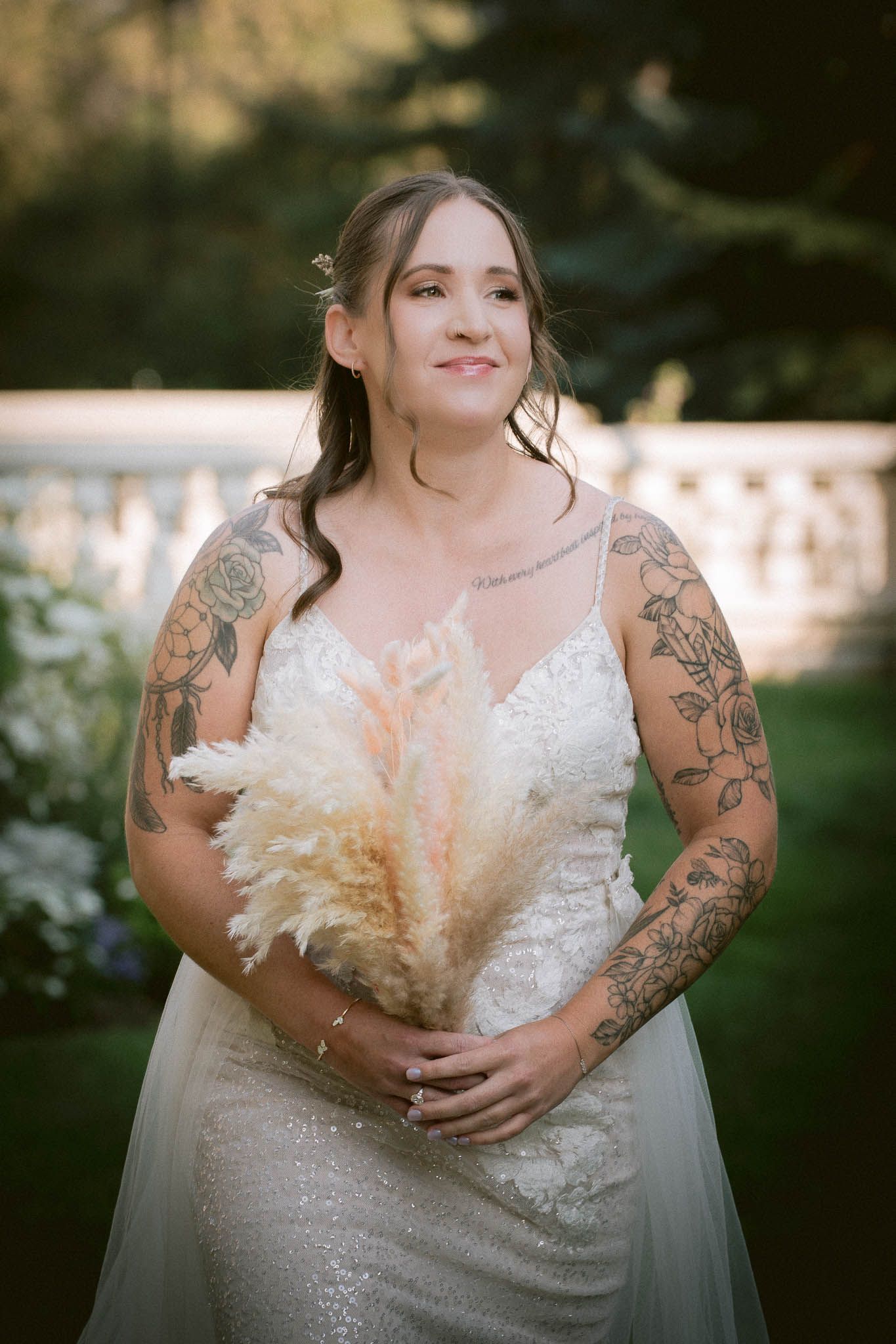 Bride in lace dress with tattoos holds dried pampas grass bouquet in outdoor setting.