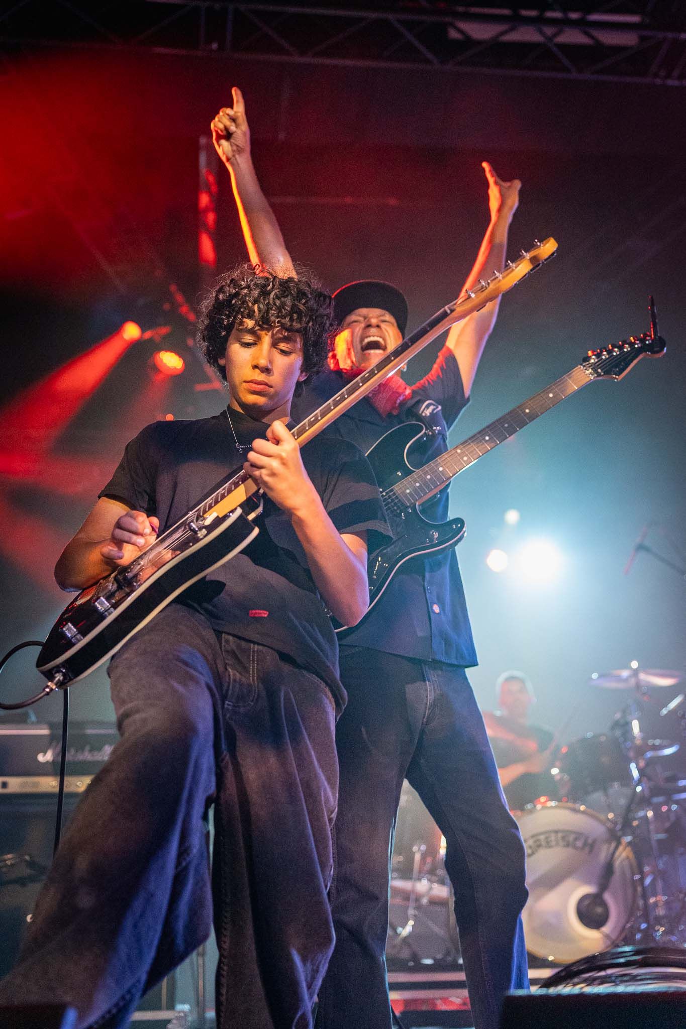 Tom Morello and son Roman Morello  perform energetically on stage with guitars under dramatic red and blue lighting during a live concert.