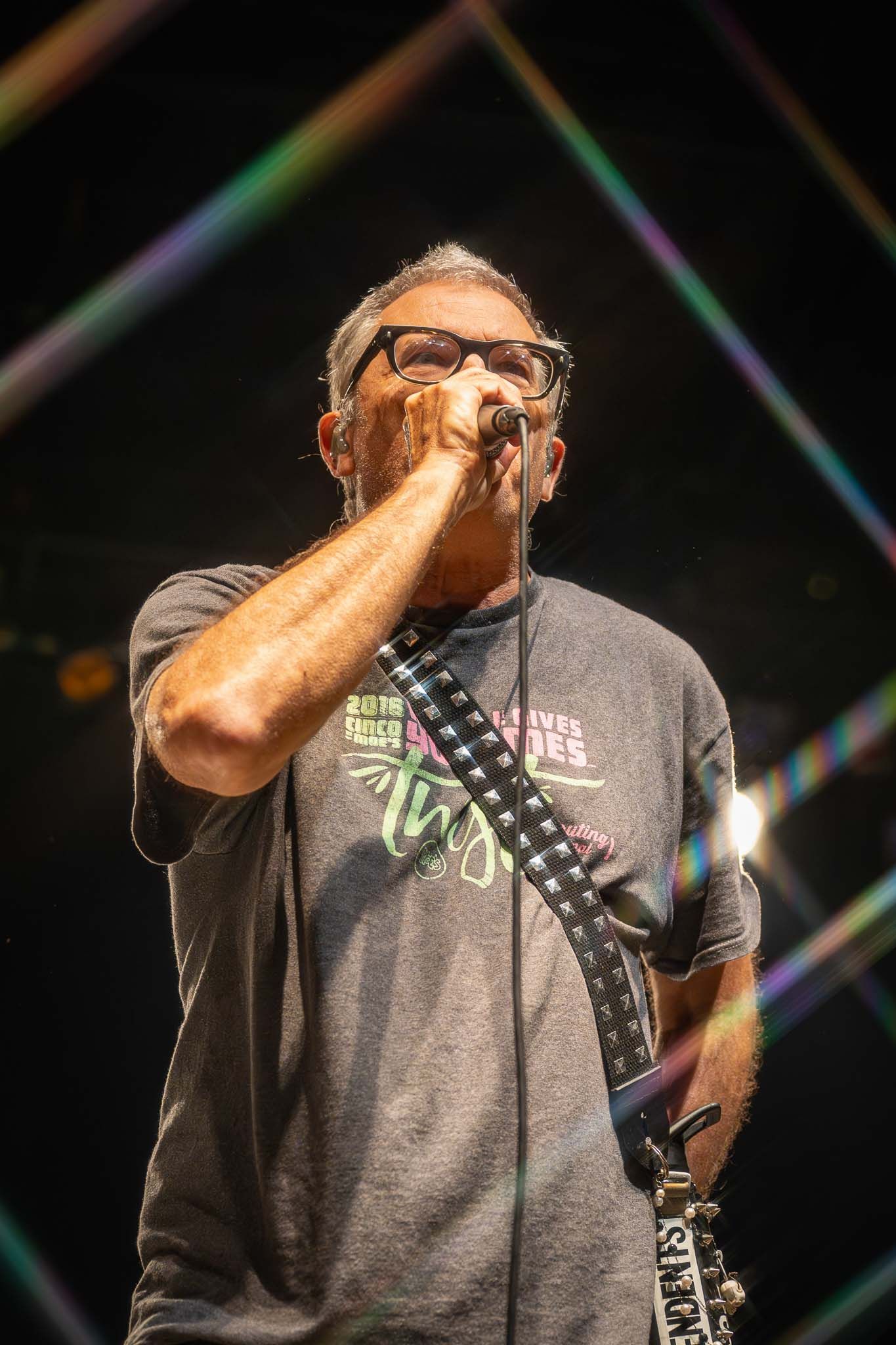 Performer in a gray t-shirt and glasses singing passionately into a microphone during a concert performance.