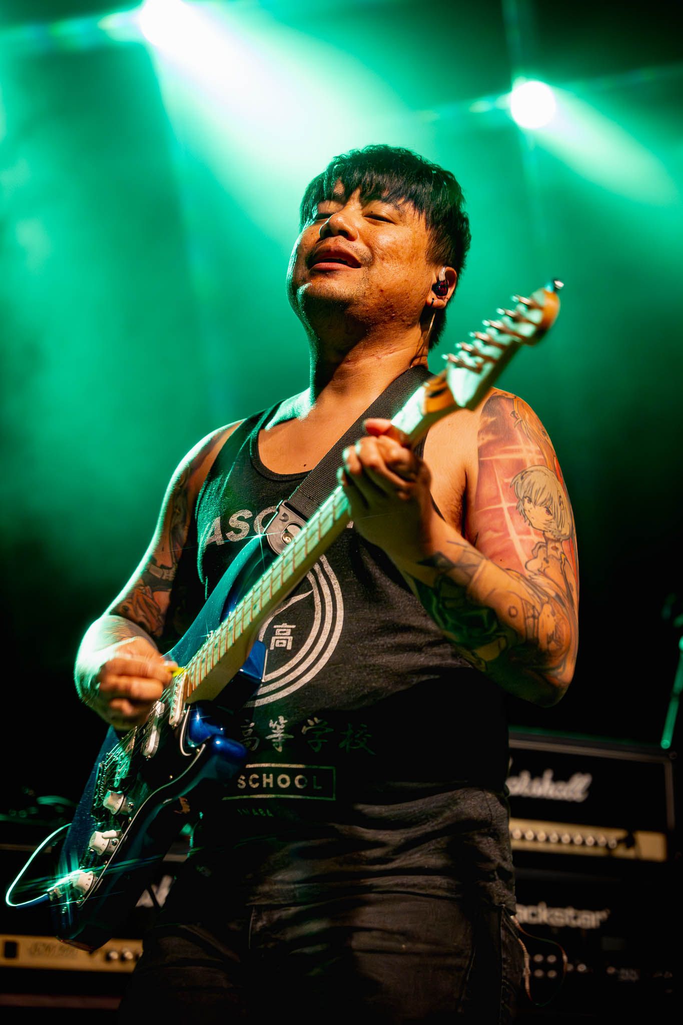Guitarist in black tank top performs energetically under green stage lighting with a blue electric guitar.